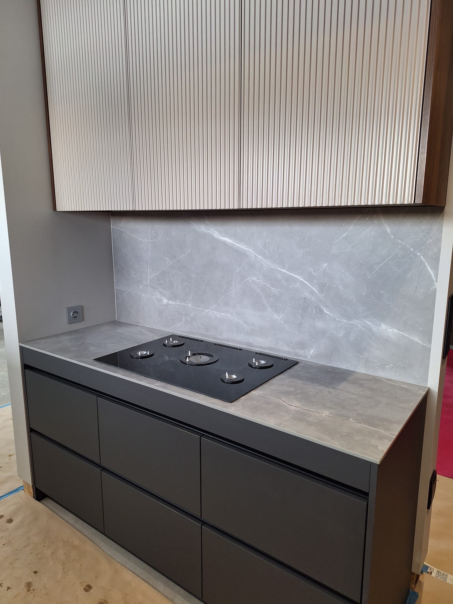 Szary blat kamienny z wbudowaną płytą grzewczą, szare szafki kuchenne z frontami bez uchwytów, ściana wykończona płytą imitującą marmur, wiszące szafki z ryflowanymi frontami.