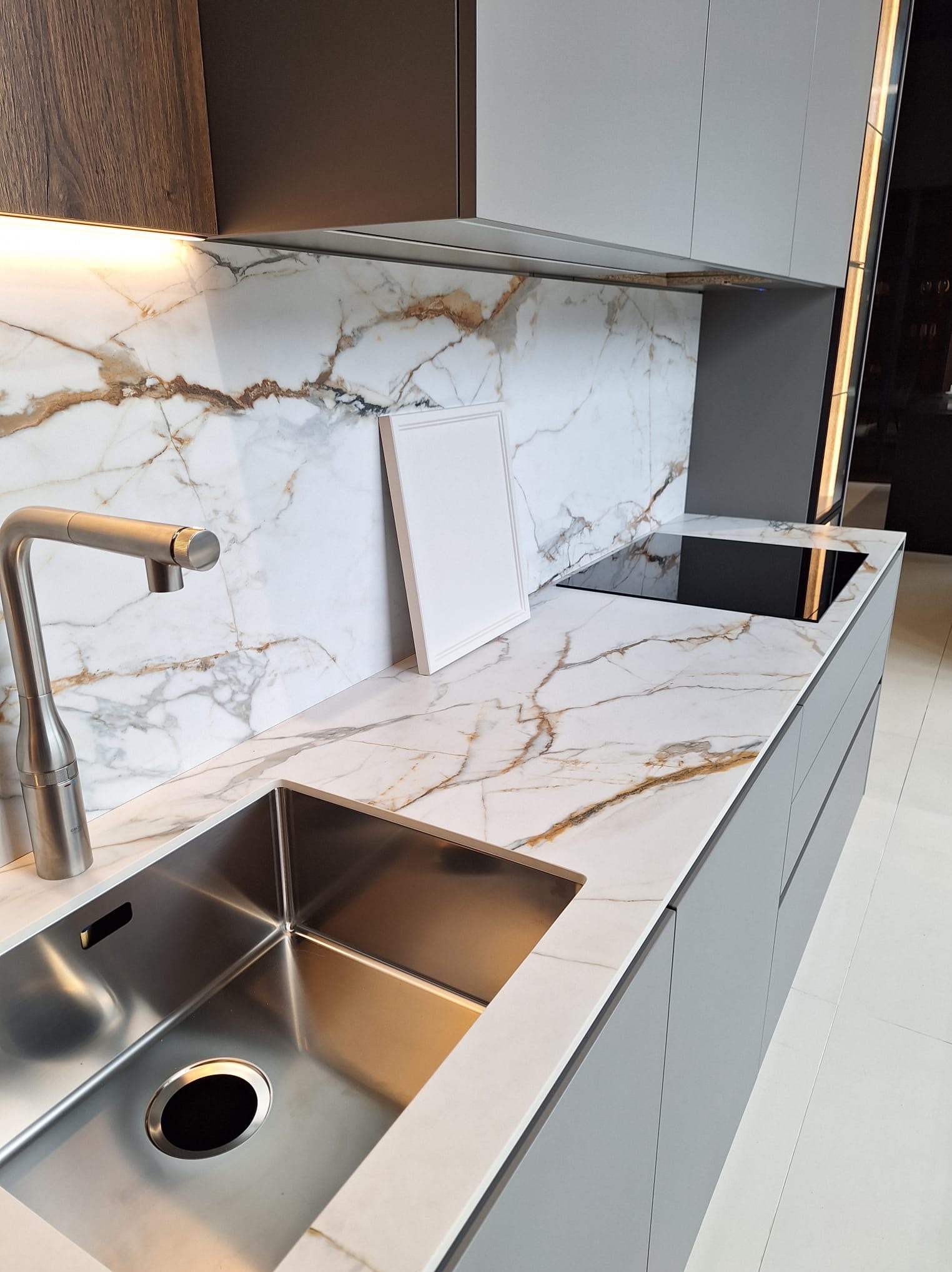 Nowoczesna kuchnia z blatem imitującym marmur, srebrnym zlewem i płytą indukcyjną, uzupełniona o białą ramkę opartą o ścianę.