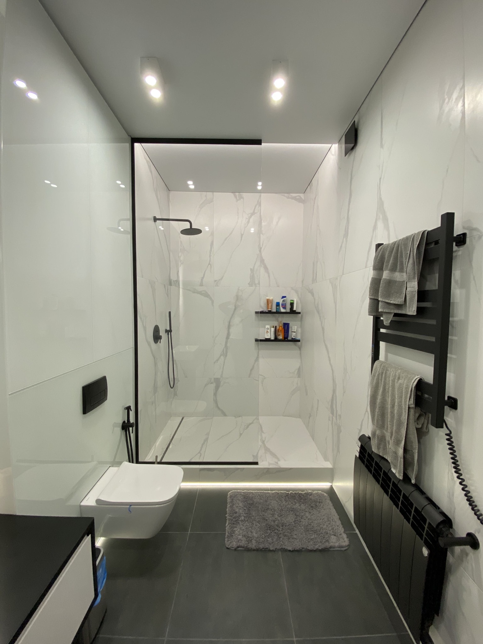 Nowoczesna łazienka z kabiną prysznicową typu walk-in, wykończona białymi płytkami imitującymi marmur, z czarnymi akcentami armatury i grzejnika drabinkowego, podświetleniem LED i wiszącą toaletą.