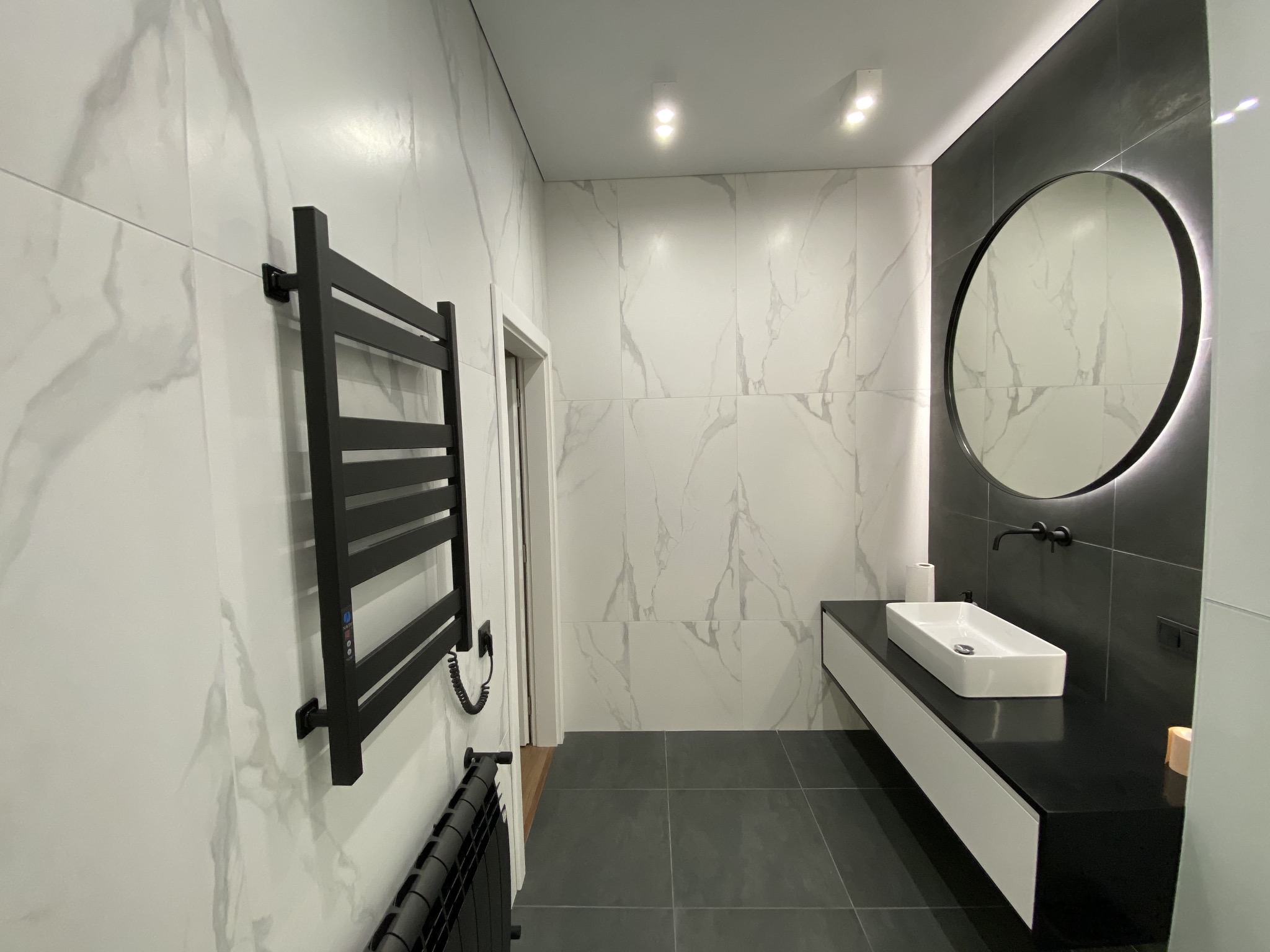 Nowoczesna łazienka z białymi marmurkowymi płytkami, czarną armaturą i okrągłym lustrem z podświetleniem LED. Widoczny grzejnik drabinkowy w kolorze czarnym.