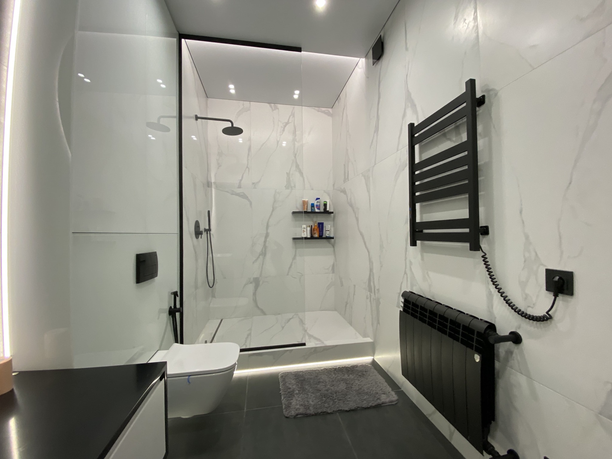 Nowoczesna łazienka z kabiną prysznicową typu walk-in, wykończona białymi płytkami imitującymi marmur, czarną armaturą i grzejnikami, podświetleniem LED i szarym dywanikiem.