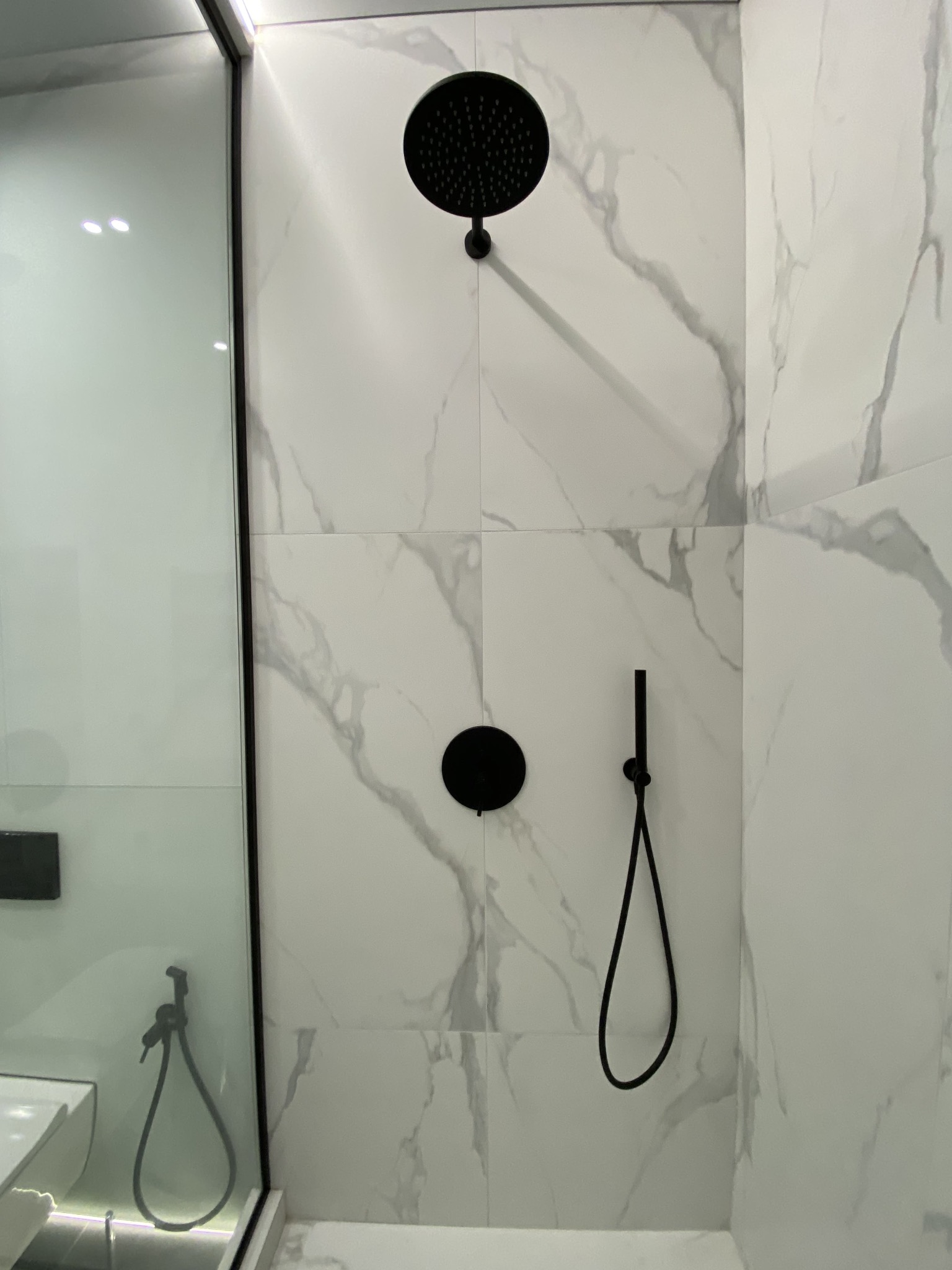 Nowoczesna kabina prysznicowa z czarną armaturą i białymi płytkami imitującymi marmur. Widoczna deszczownica, słuchawka prysznicowa i szklana ściana kabiny.