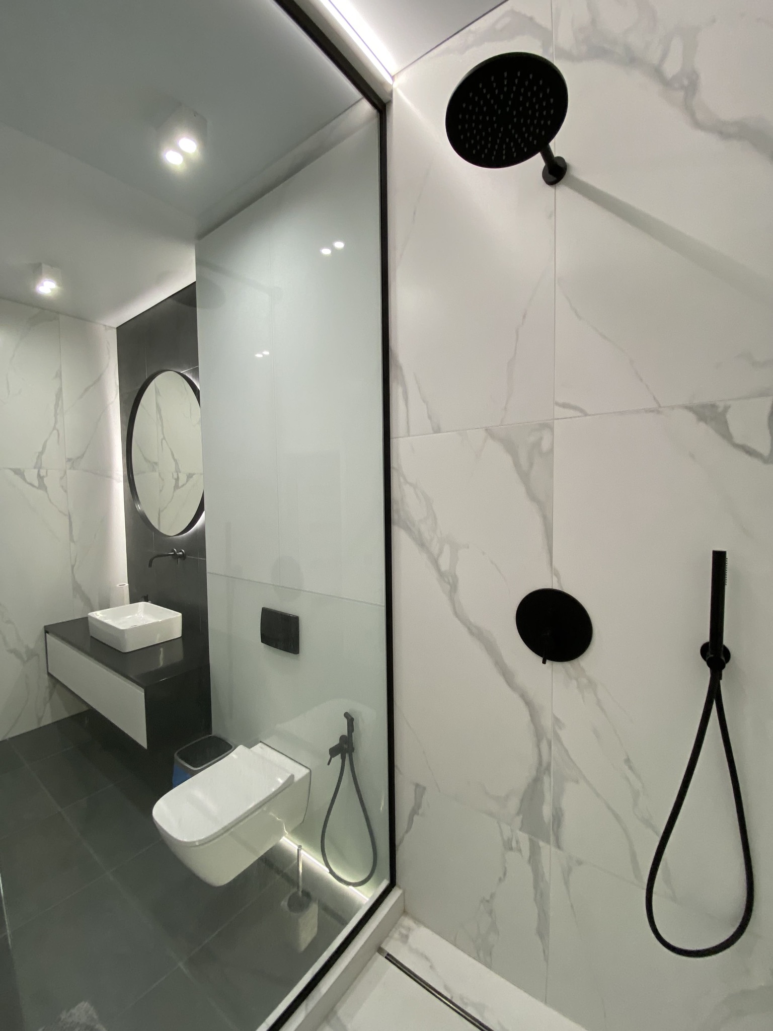 Nowoczesna łazienka z białymi marmurowymi płytkami, czarną armaturą, podwieszaną toaletą i okrągłym lustrem z podświetleniem LED.