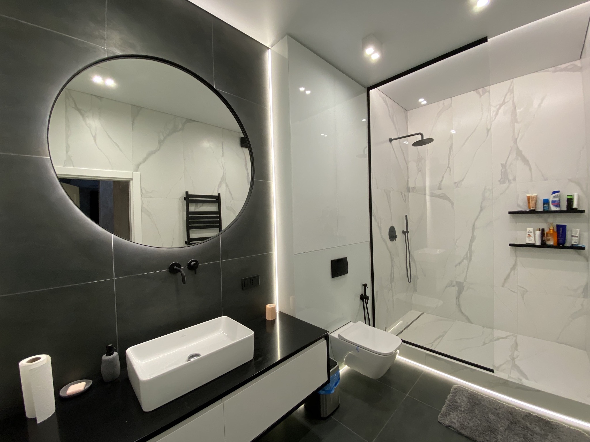 Nowoczesna łazienka w czarno-białej kolorystyce z okrągłym lustrem, podświetleniem LED, wiszącą toaletą i kabiną prysznicową typu walk-in z marmurowymi płytkami.