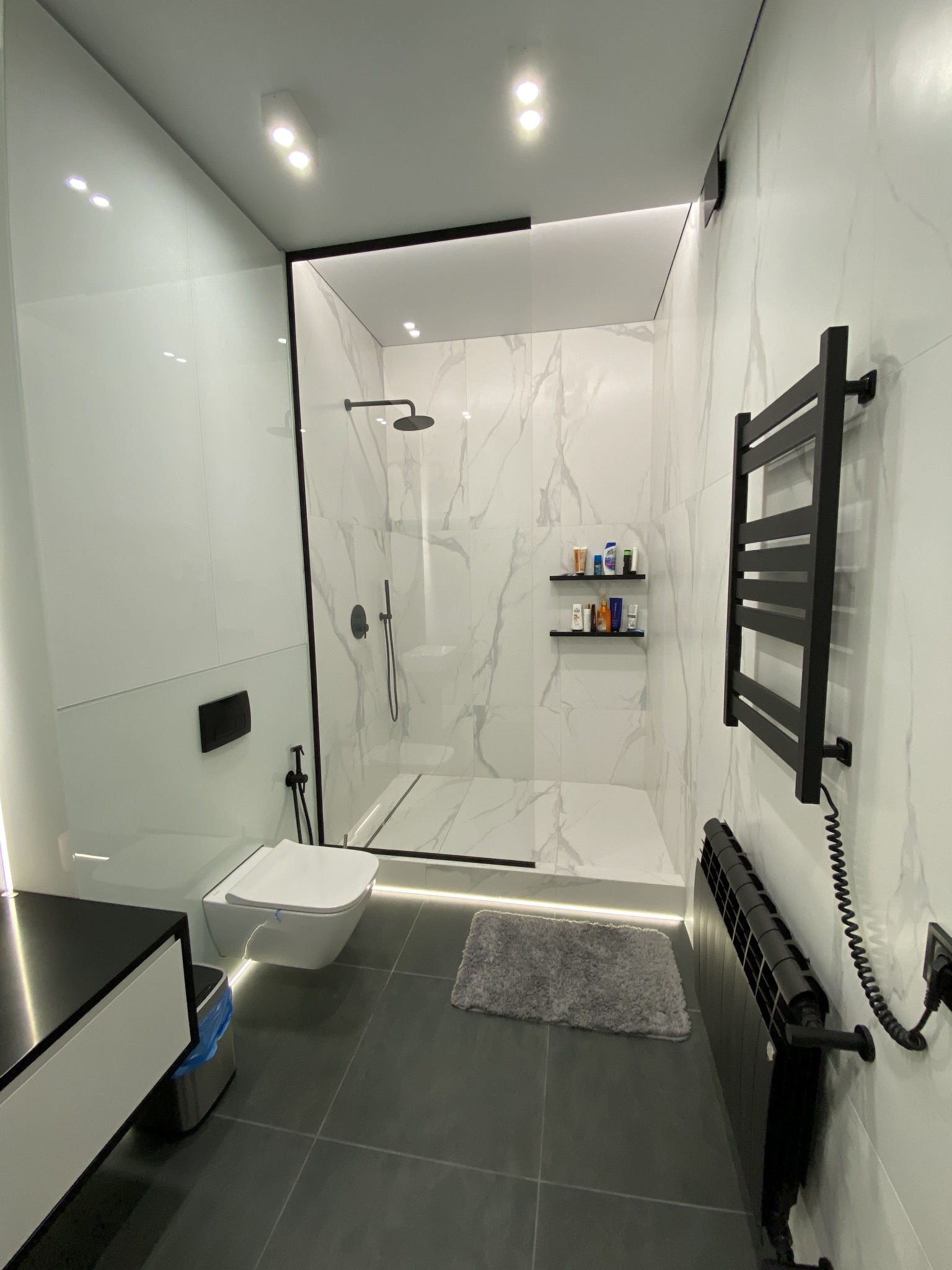 Nowoczesna łazienka z kabiną prysznicową typu walk-in, białe marmurowe płytki, czarne akcenty armatury i grzejników, podświetlenie LED pod toaletą i kabiną, szary dywanik.
