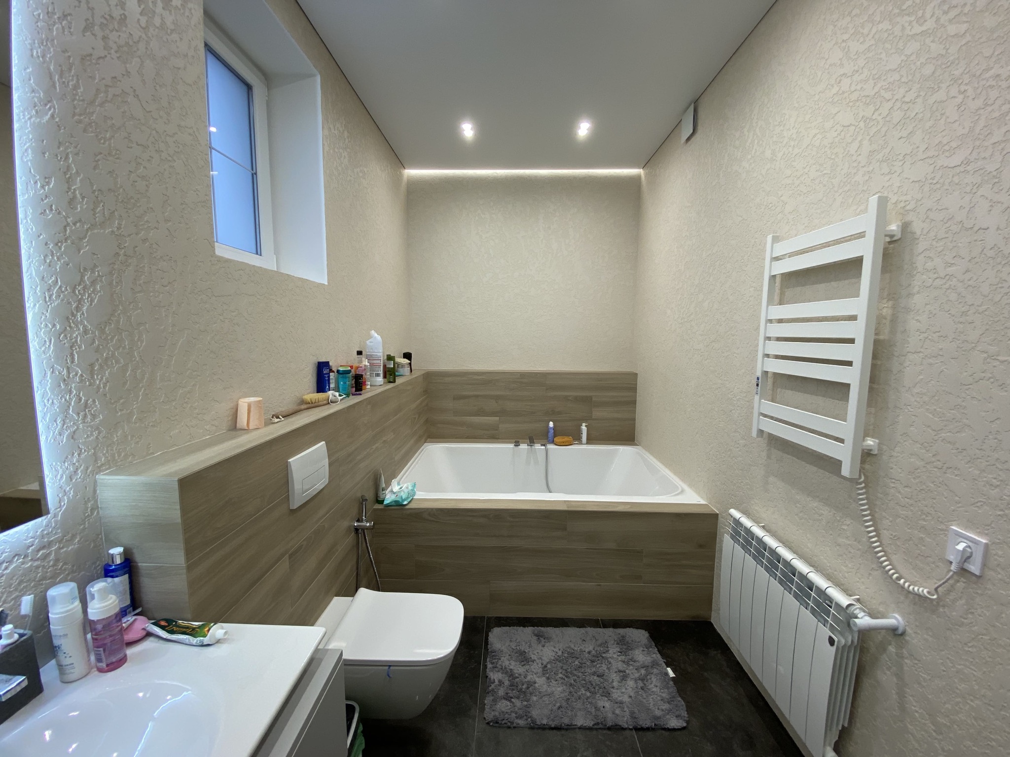 Jasna łazienka z wanną obudowaną drewnopodobnymi płytkami, toaletą, grzejnikiem drabinkowym i ściennym, oraz teksturowanymi ścianami w odcieniach beżu.