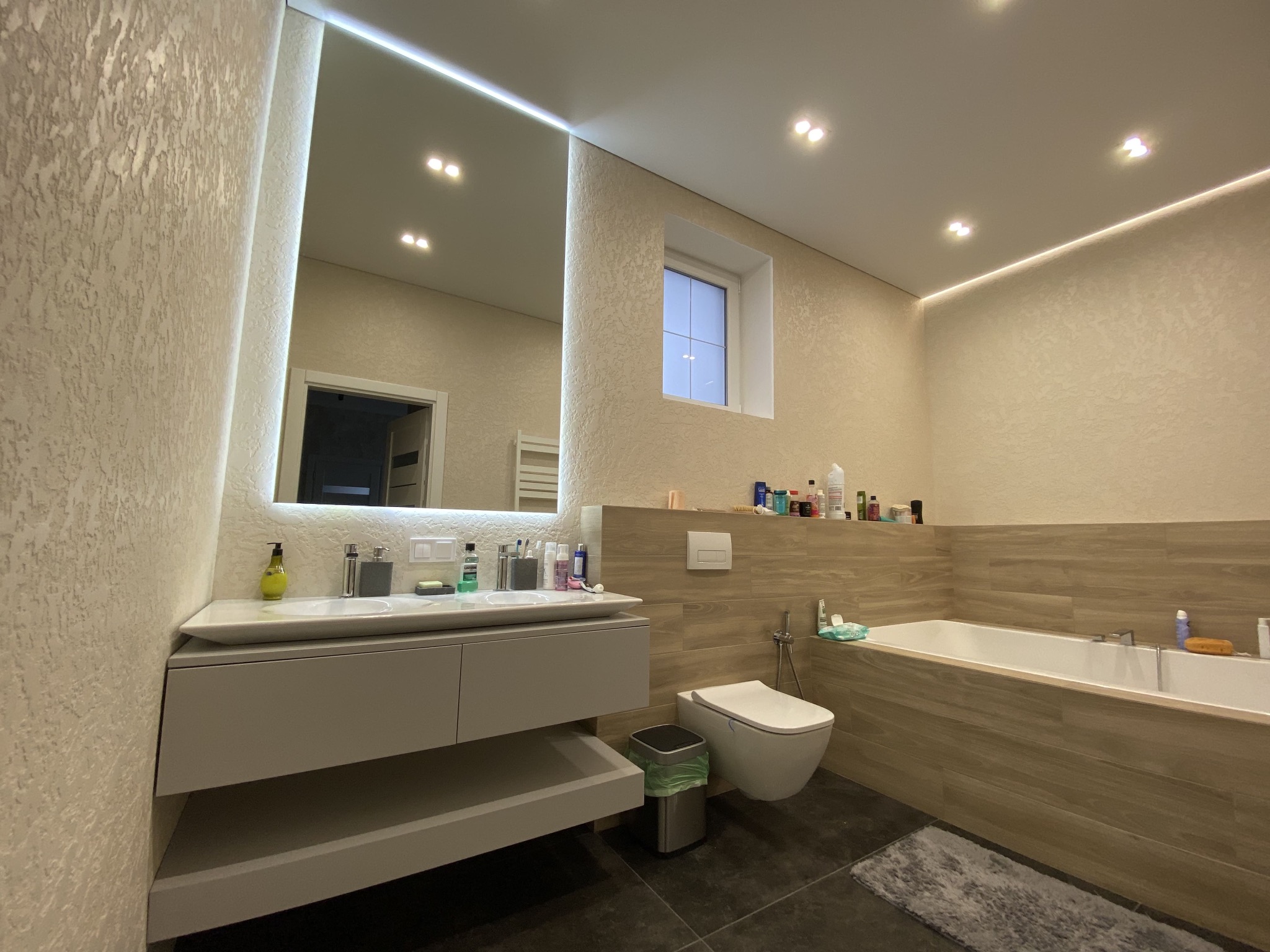 Nowoczesna łazienka w Warszawie z podświetlanym lustrem, wiszącą umywalką i wanną obudowaną płytkami imitującymi drewno, uzupełniona o minimalistyczną toaletę i kosz na śmieci.