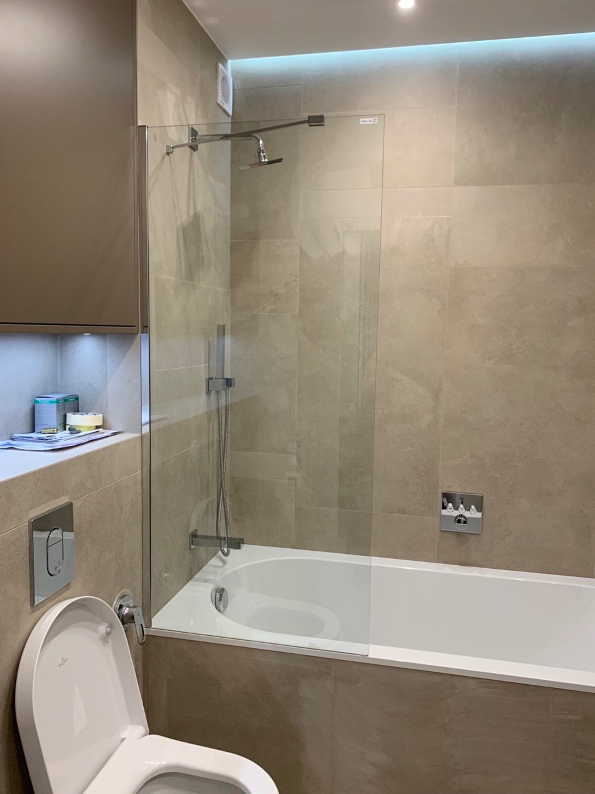 Nowoczesna łazienka z beżowymi płytkami, białą wanną z szklanym parawanem, podświetlanym sufitem LED i zabudową meblową z oświetleniem punktowym nad toaletą.