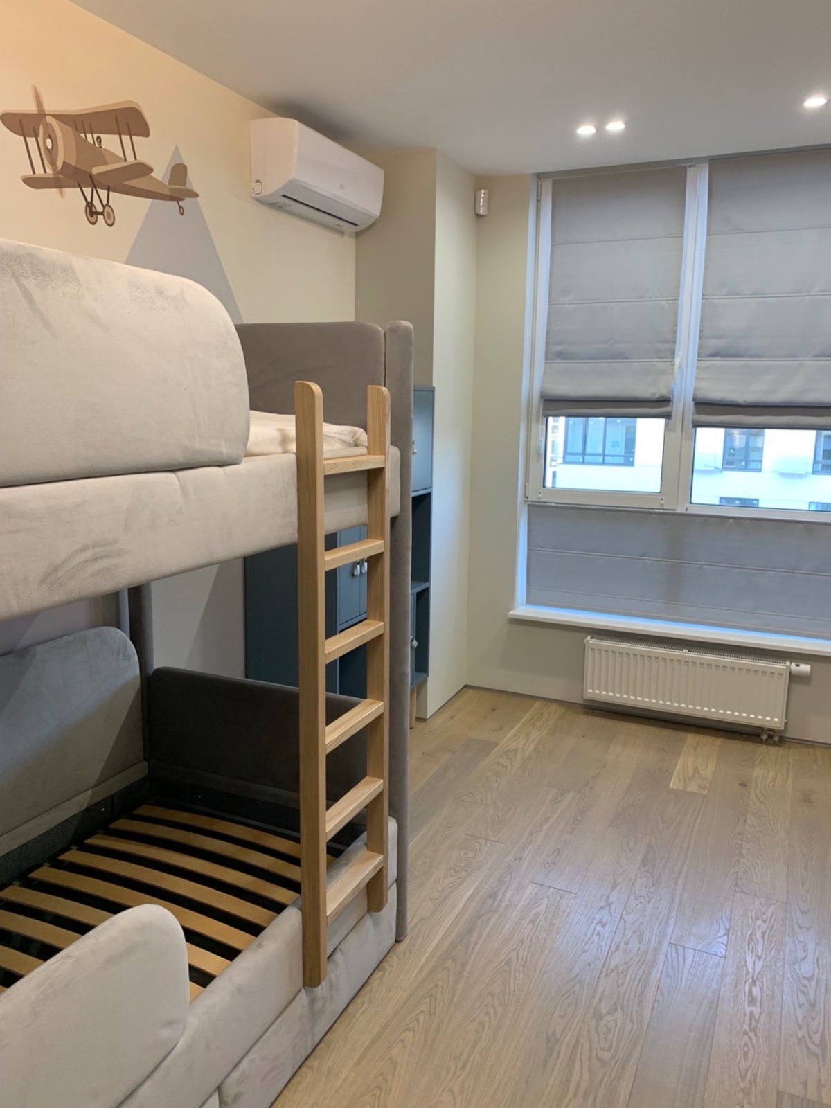 Pokój dziecięcy z szarym łóżkiem piętrowym, drewnianą drabinką, rysunkiem samolotu na ścianie, klimatyzatorem i oknem z szarymi roletami rzymskimi.