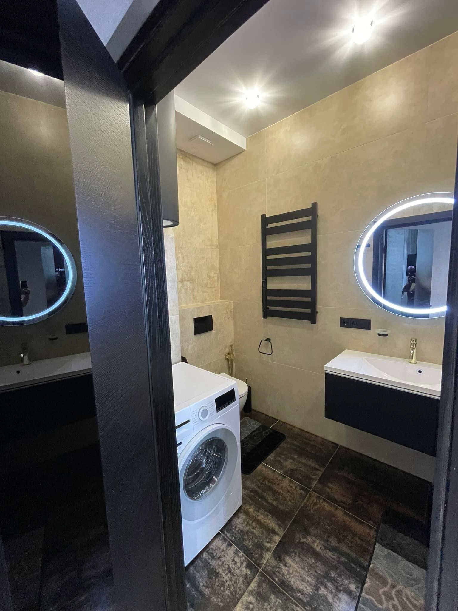 Nowoczesna łazienka z beżowymi płytkami, okrągłym lustrem z podświetleniem LED, czarną szafką pod umywalką, grzejnikiem drabinkowym i pralką, widziana z perspektywy wejścia przez czarną futrynę.