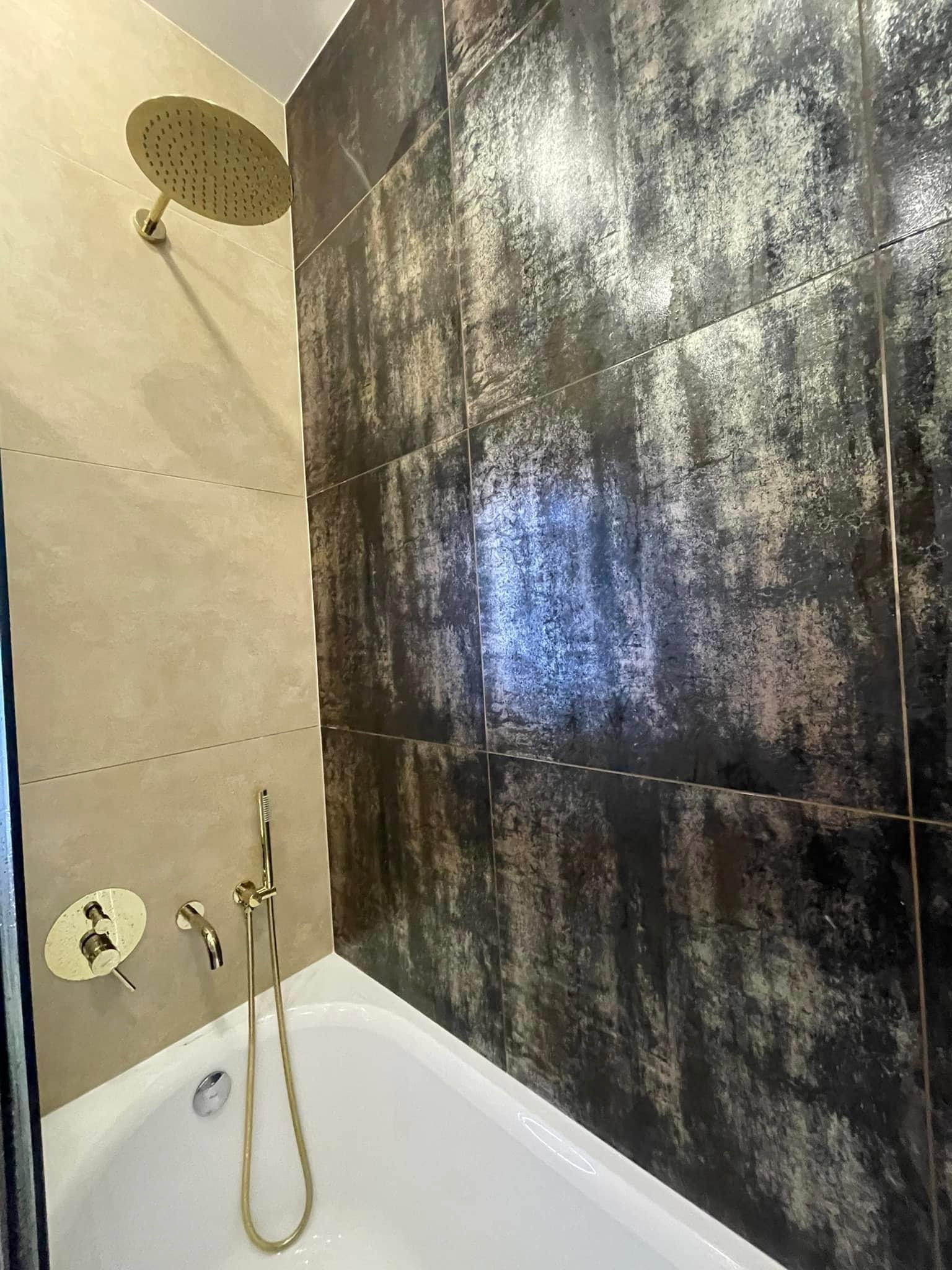 Nowoczesna łazienka z wanną, złotą armaturą i ciemnymi płytkami imitującymi kamień na ścianie.