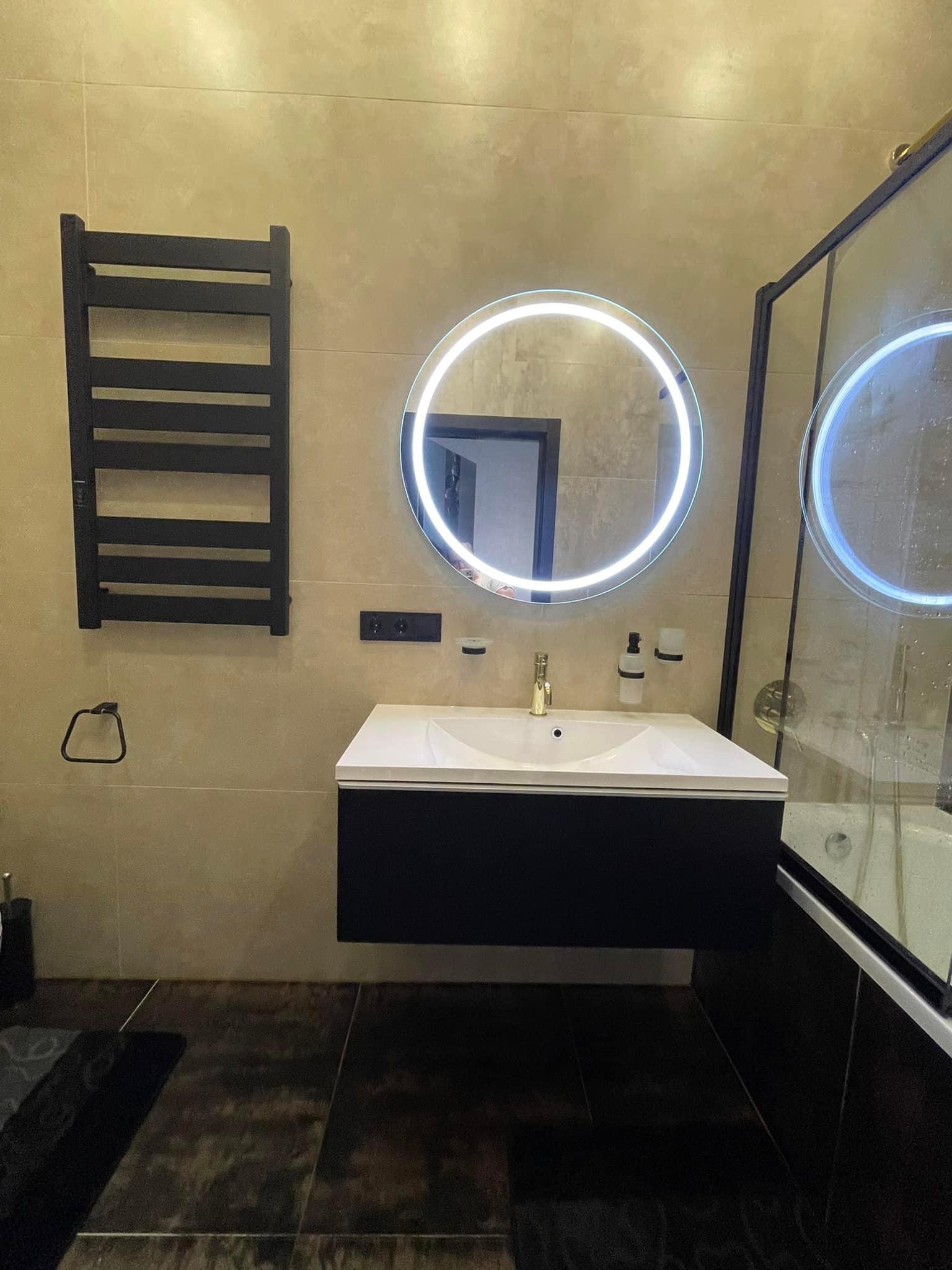 Nowoczesna łazienka z beżowymi płytkami, czarną armaturą i okrągłym lustrem z podświetleniem LED. Widoczna umywalka nablatowa z czarną szafką, grzejnik drabinkowy i kabina prysznicowa z czarnymi...