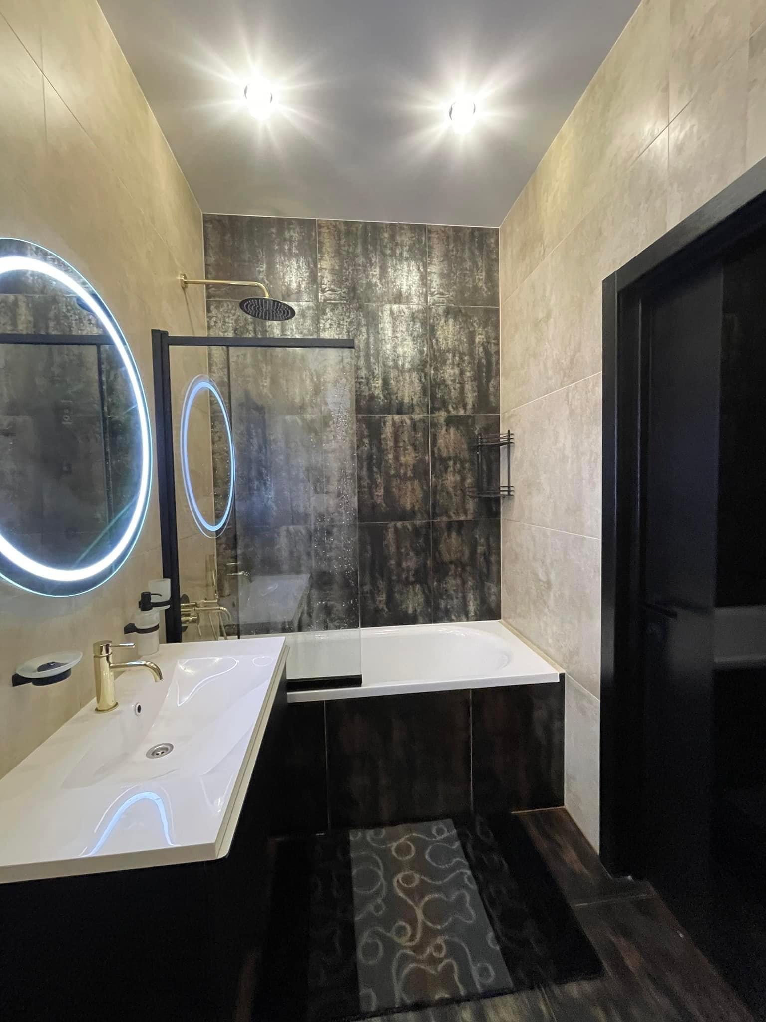 Nowoczesna łazienka z podświetlanym lustrem, złotą armaturą, wanną obudowaną ciemnymi płytkami i dekoracyjnym dywanikiem.