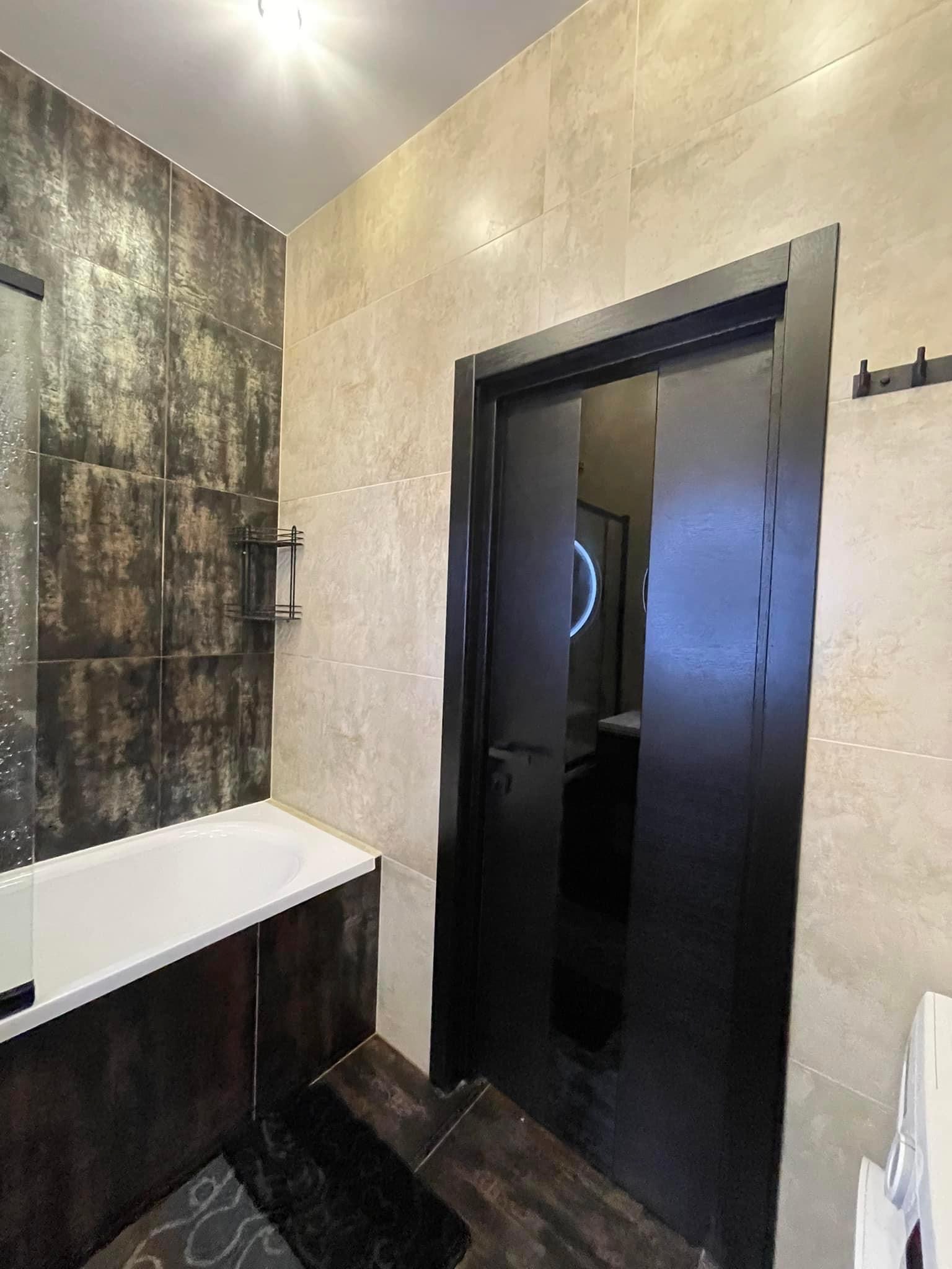 Nowoczesna łazienka z ciemnymi płytkami imitującymi metal, jasnymi płytkami imitującymi kamień, czarną ramą drzwi przesuwnych i widoczną wanną z parawanem.