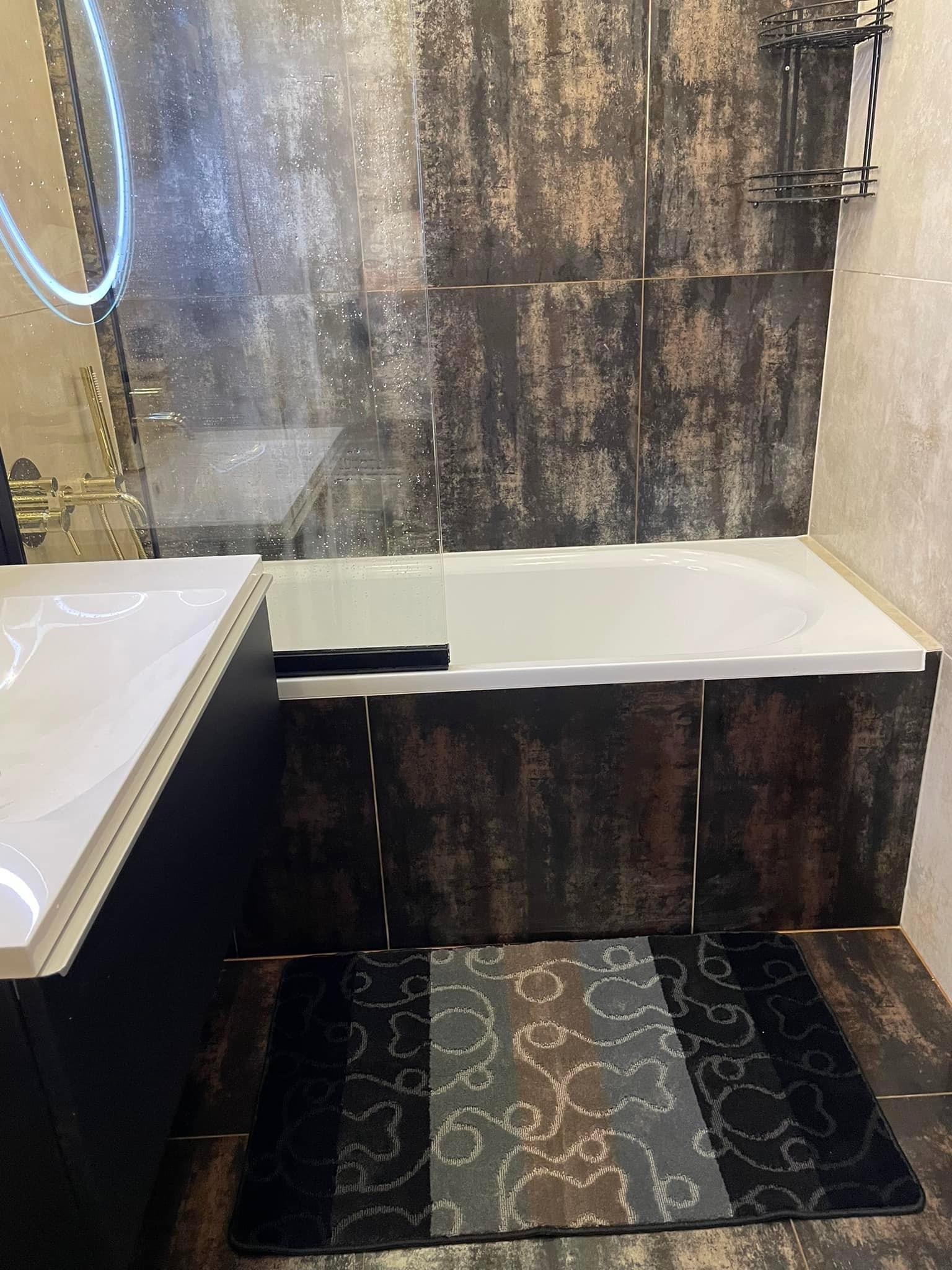 Nowoczesna łazienka z wanną obudowaną panelami imitującymi ciemne drewno, brązowo-szare płytki na ścianach i podłodze, szklany parawan nawannowy oraz dywanik w odcieniach szarości i czerni.