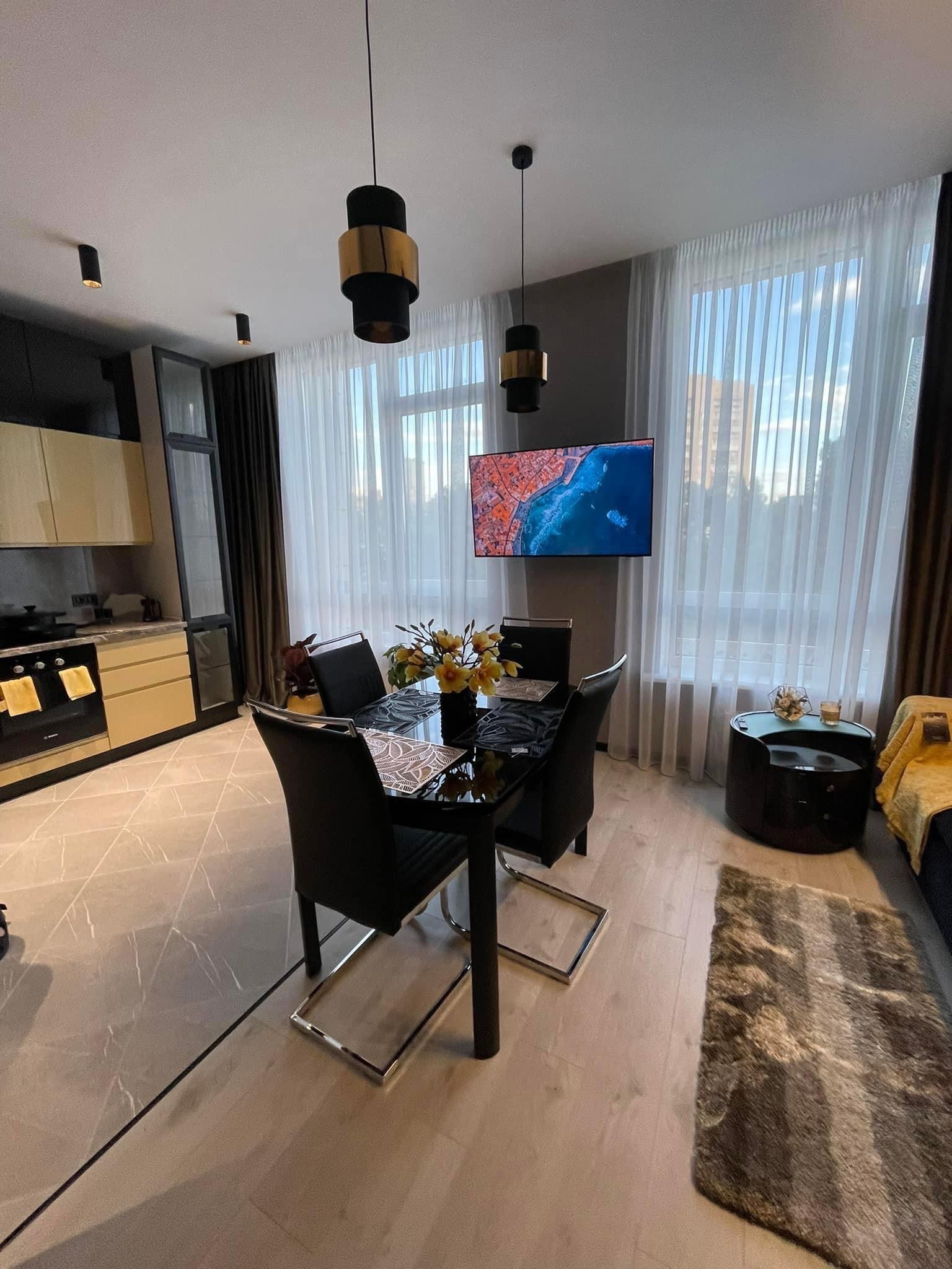 Nowoczesne wnętrze jadalni z czarnym stołem i krzesłami na tle kuchni z beżowymi szafkami, telewizor na ścianie i jasne zasłony w oknach.