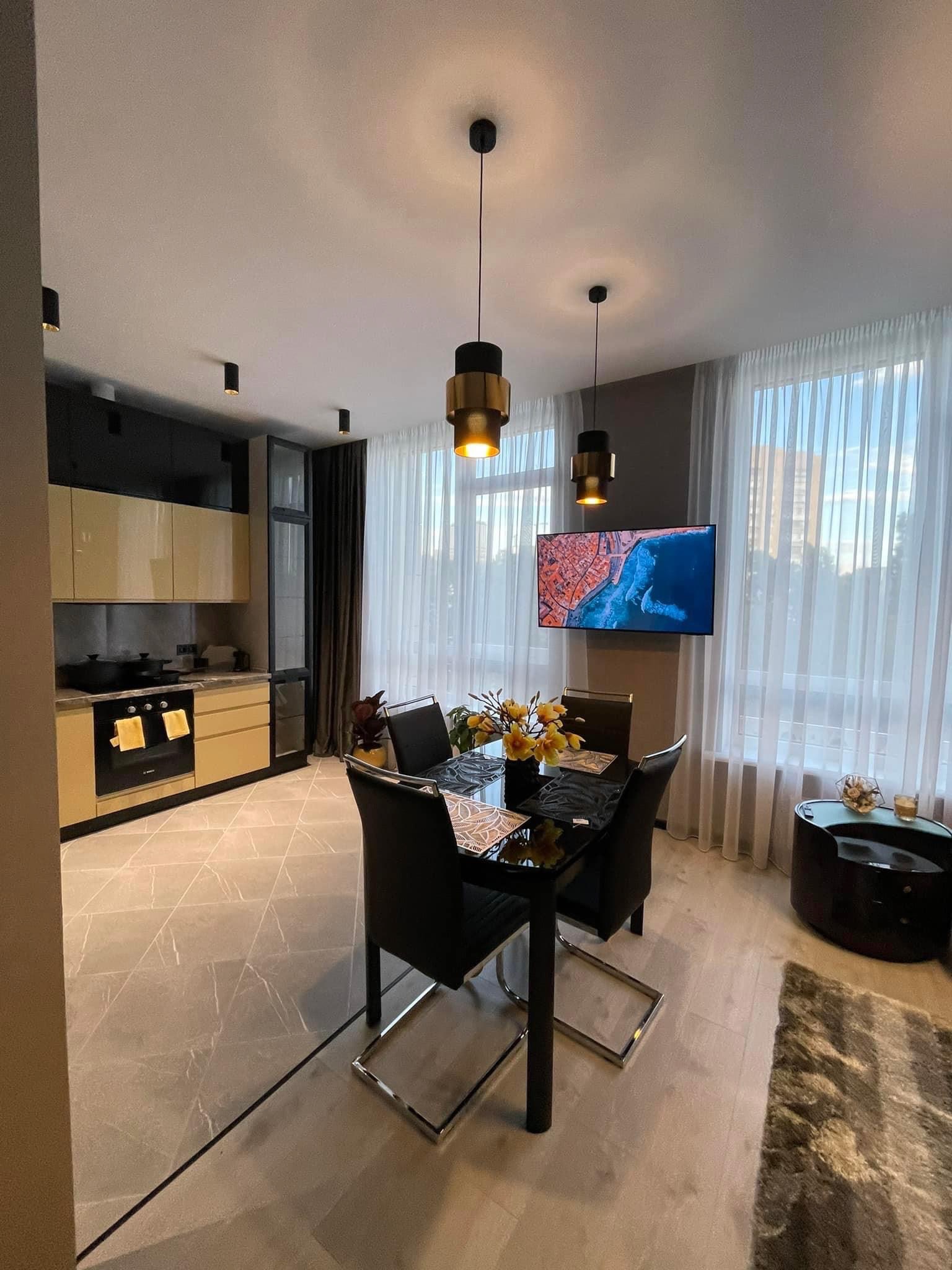 Nowoczesna jadalnia z czarnym stołem i krzesłami, żółtymi kwiatami, telewizorem na ścianie i aneksem kuchennym z beżowo-czarnymi szafkami.