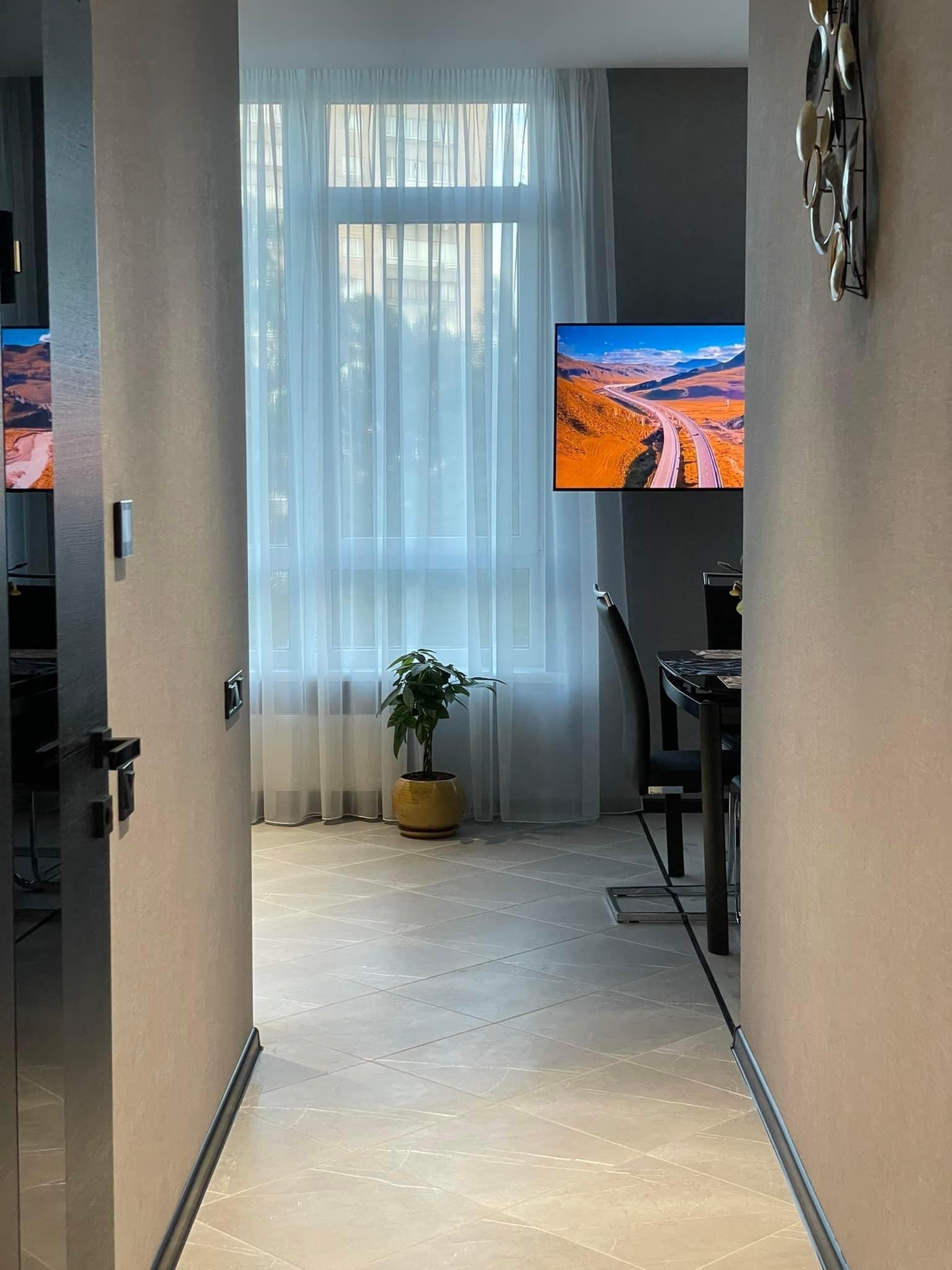 Wnętrze mieszkania z widokiem na salon: szare ściany, jasna podłoga z płytek, okno z białymi zasłonami, telewizor z krajobrazem i dekoracja ścienna z metalowych kół.