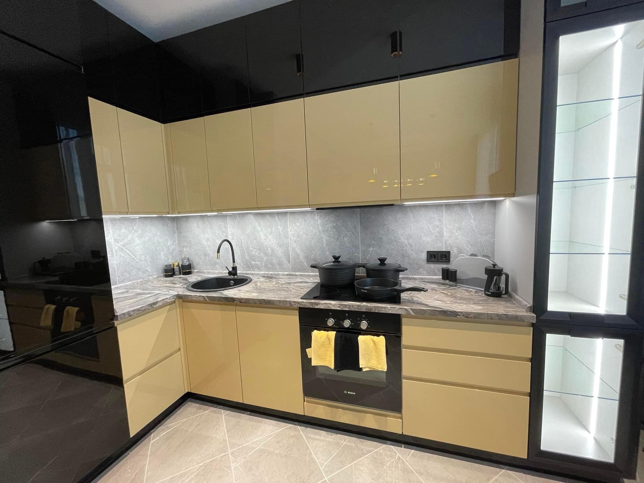 Nowoczesna kuchnia z beżowymi szafkami na wysoki połysk, czarnymi elementami górnymi i marmurowym blatem, oświetlona listwami LED pod szafkami wiszącymi.