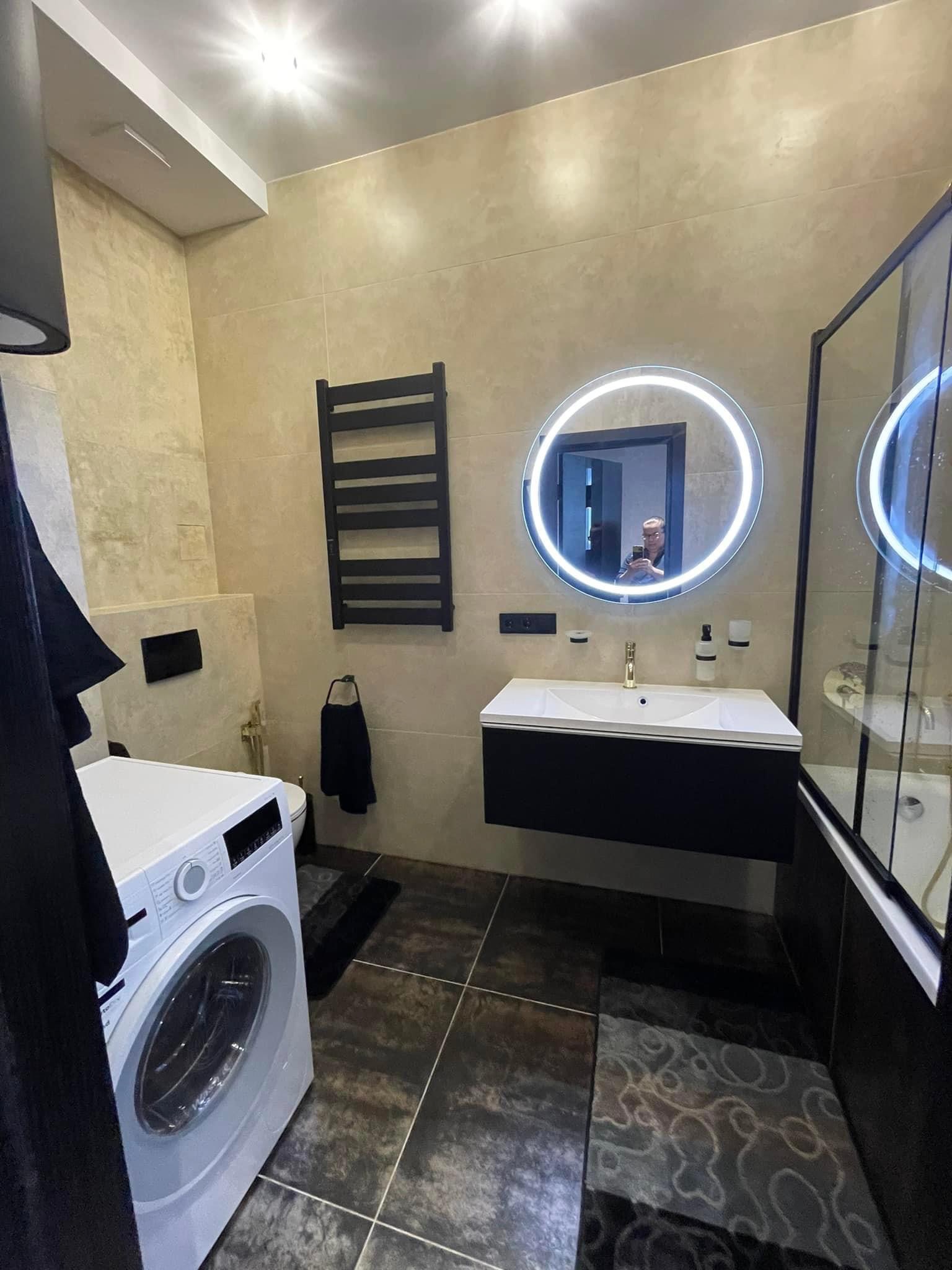 Nowoczesna łazienka z beżowymi płytkami, podświetlanym okrągłym lustrem, czarną szafką pod umywalką i pralką, widoczna osoba robiąca zdjęcie w lustrze.