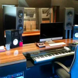 Profesjonalne stanowisko odsłuchowe w studiu z monitorami bliskiego i średniego pola, klawiaturą sterującą, komputerem iMac i butelką wody na lakierowanym biurku.