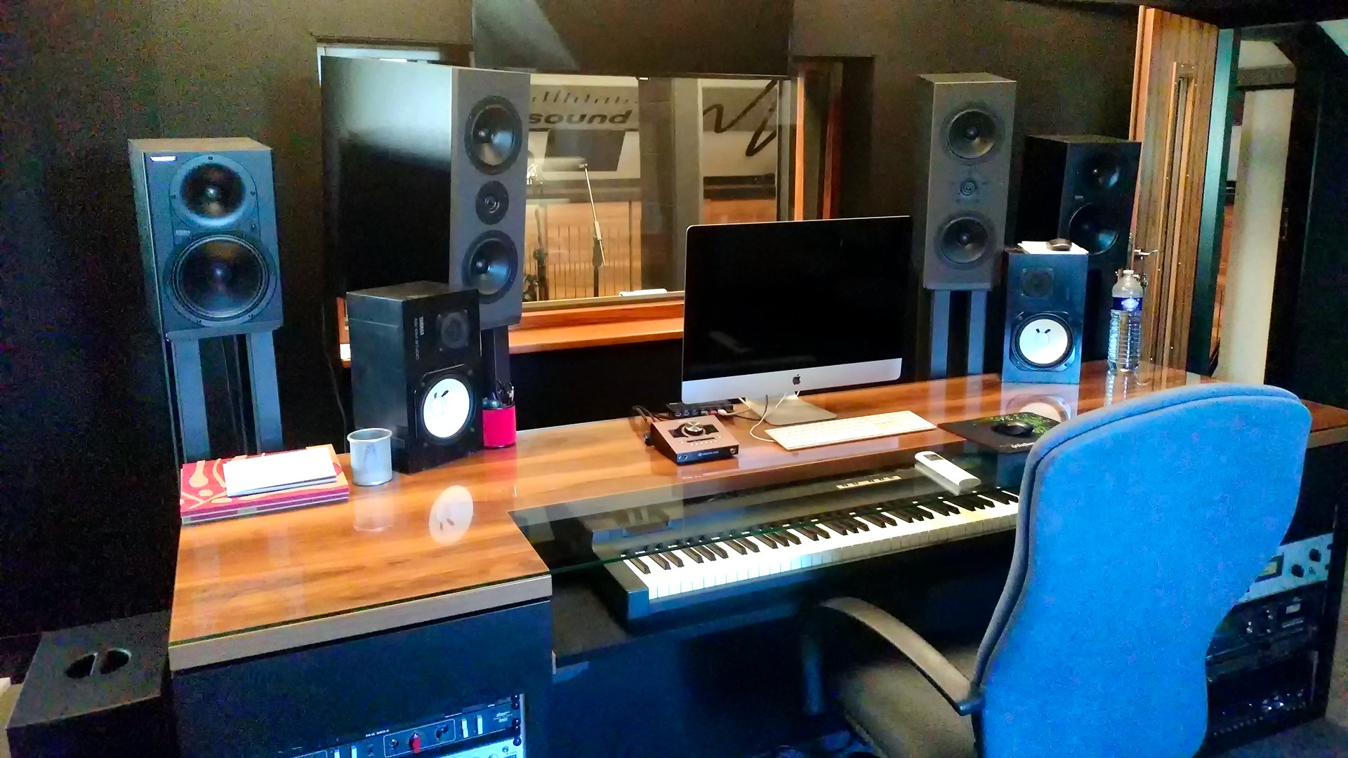 Profesjonalne stanowisko odsłuchowe w studiu z monitorami bliskiego i średniego pola, klawiaturą sterującą, komputerem iMac i butelką wody na lakierowanym biurku.