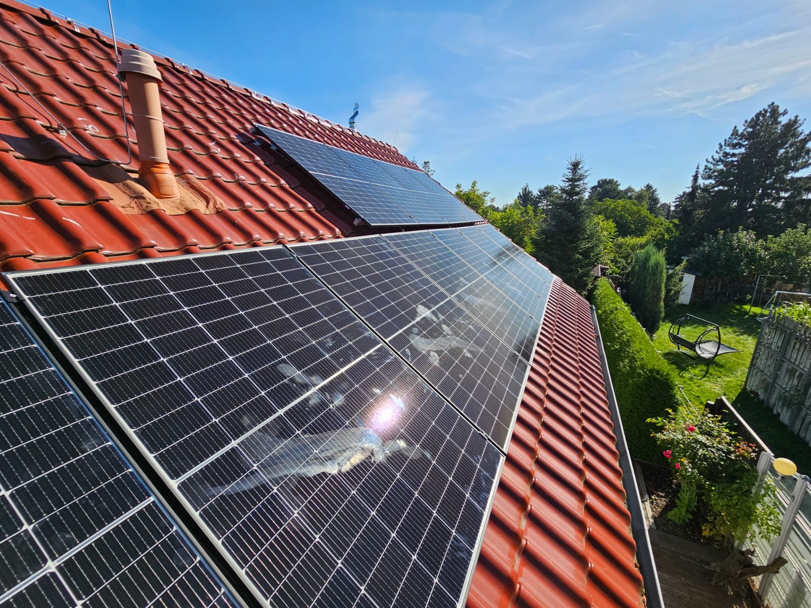 Panele słoneczne zamontowane na czerwonym dachu z dachówki ceramicznej, widoczne odbicie nieba i chmur na powierzchni paneli, w tle zieleń drzew i krzewów.