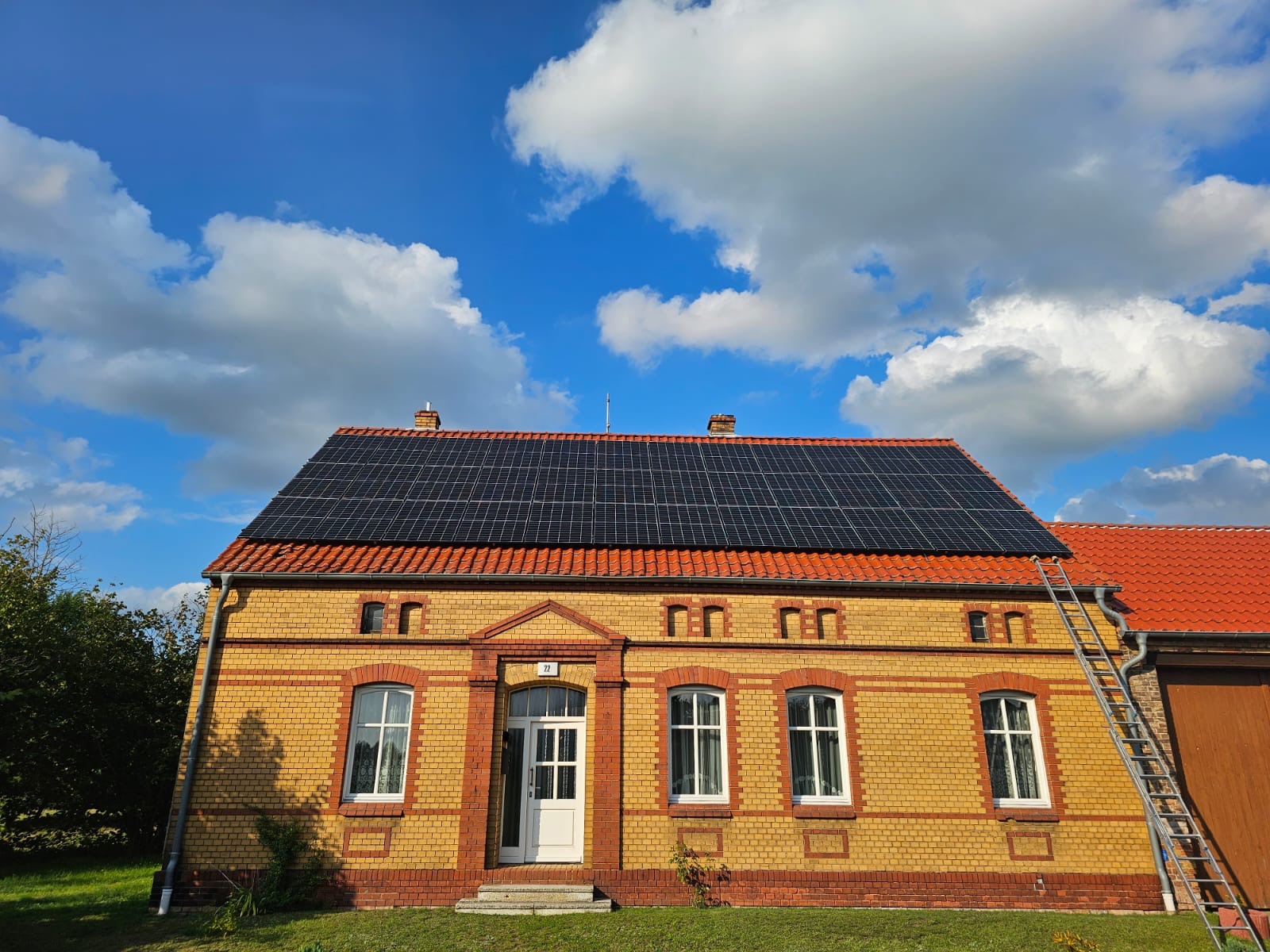 Żółty, ceglany dom z czerwoną dachówką, na której zamontowane są czarne panele słoneczne; drabina oparta o dach. Błękitne niebo z białymi chmurami w tle.