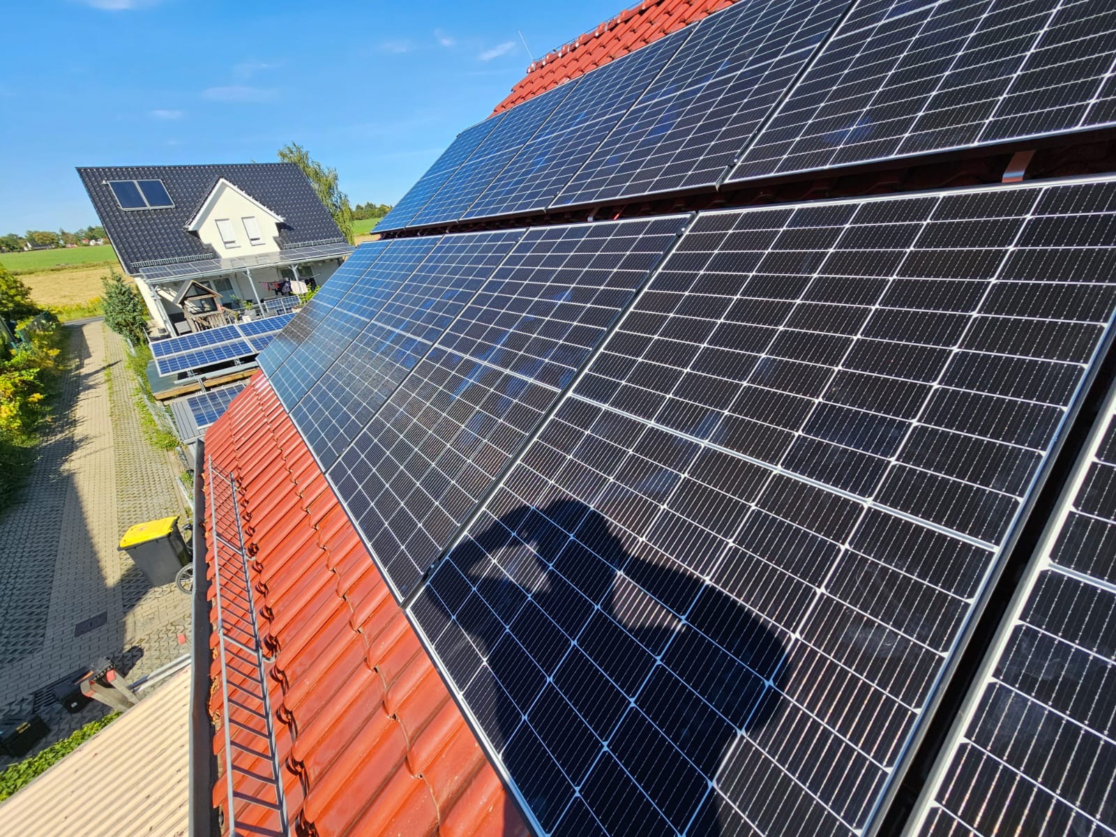 Panele słoneczne na czerwonym, dachówkowym dachu domu jednorodzinnego w słoneczny dzień, z widocznym cieniem osoby instalującej.