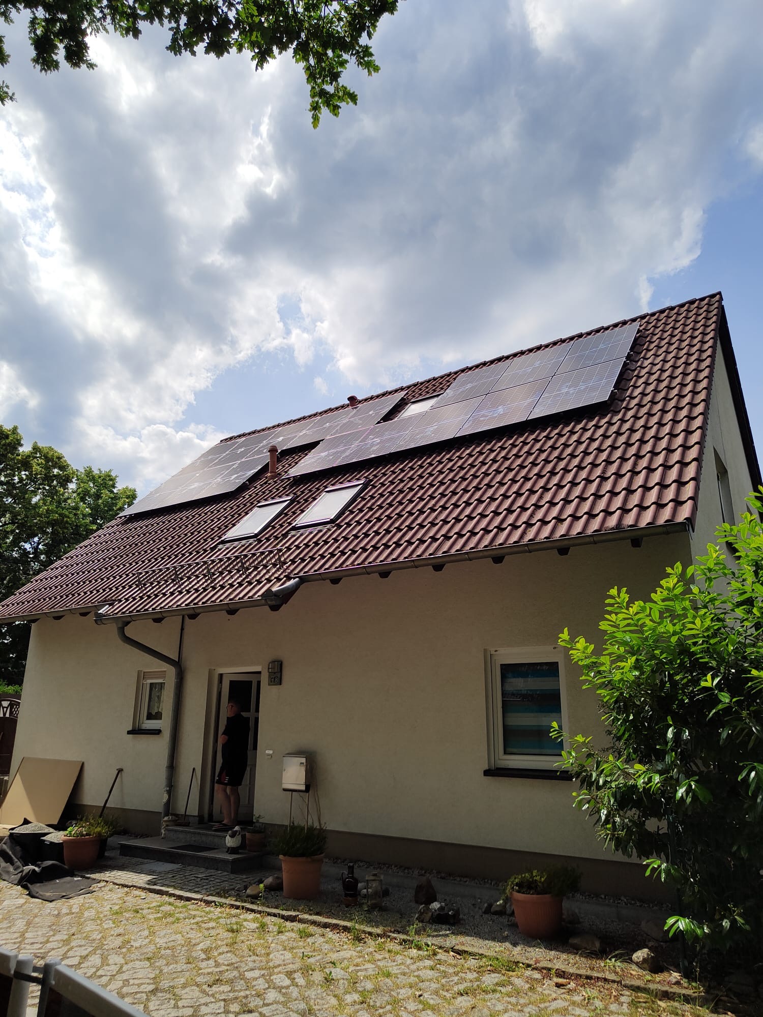 Dom jednorodzinny z panelami słonecznymi na brązowym dachu, widoczne okna dachowe i osoba stojąca w drzwiach wejściowych.