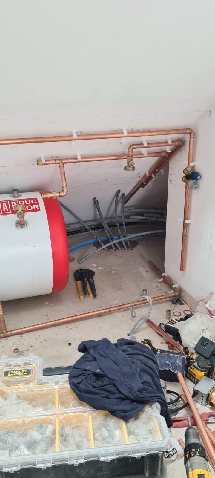 Montaż instalacji centralnego ogrzewania z miedzianych rur, widoczny zasobnik wody, narzędzia i skrzynka Stanley Fatmax z elementami instalacyjnymi, surowe ściany i podłoga.