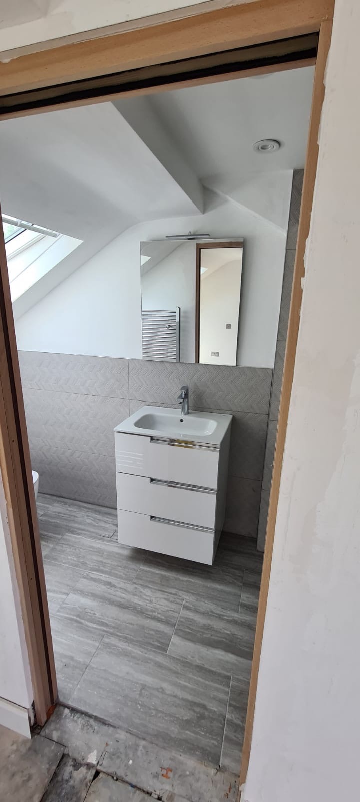 Nowoczesna łazienka na poddaszu z białą szafką pod umywalką, szarą glazurą i lustrem. Widoczny fragment okna dachowego i niedokończone wykończenie ościeżnicy.
