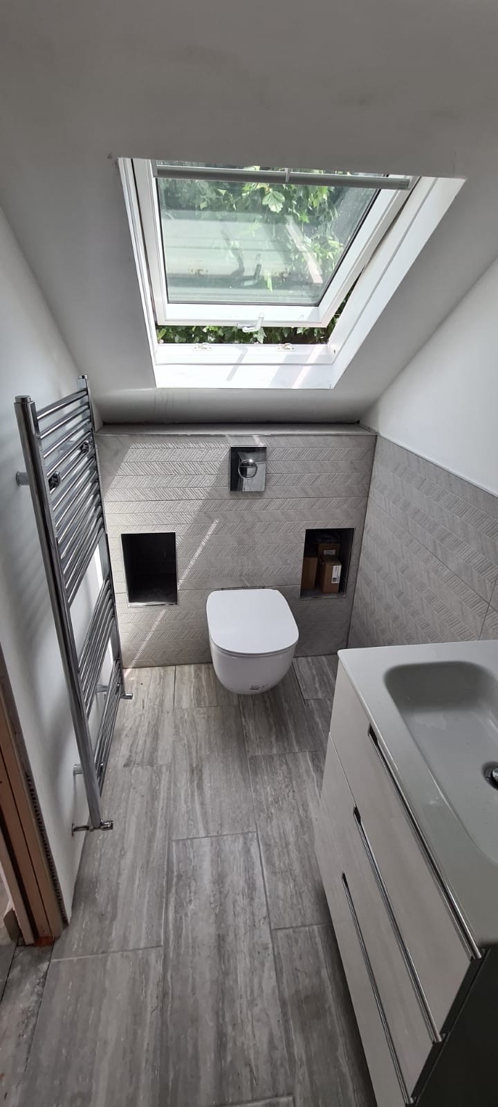 Nowoczesna łazienka na poddaszu z oknem dachowym, szare płytki w jodełkę na ścianie, wisząca toaleta i grzejnik drabinkowy.