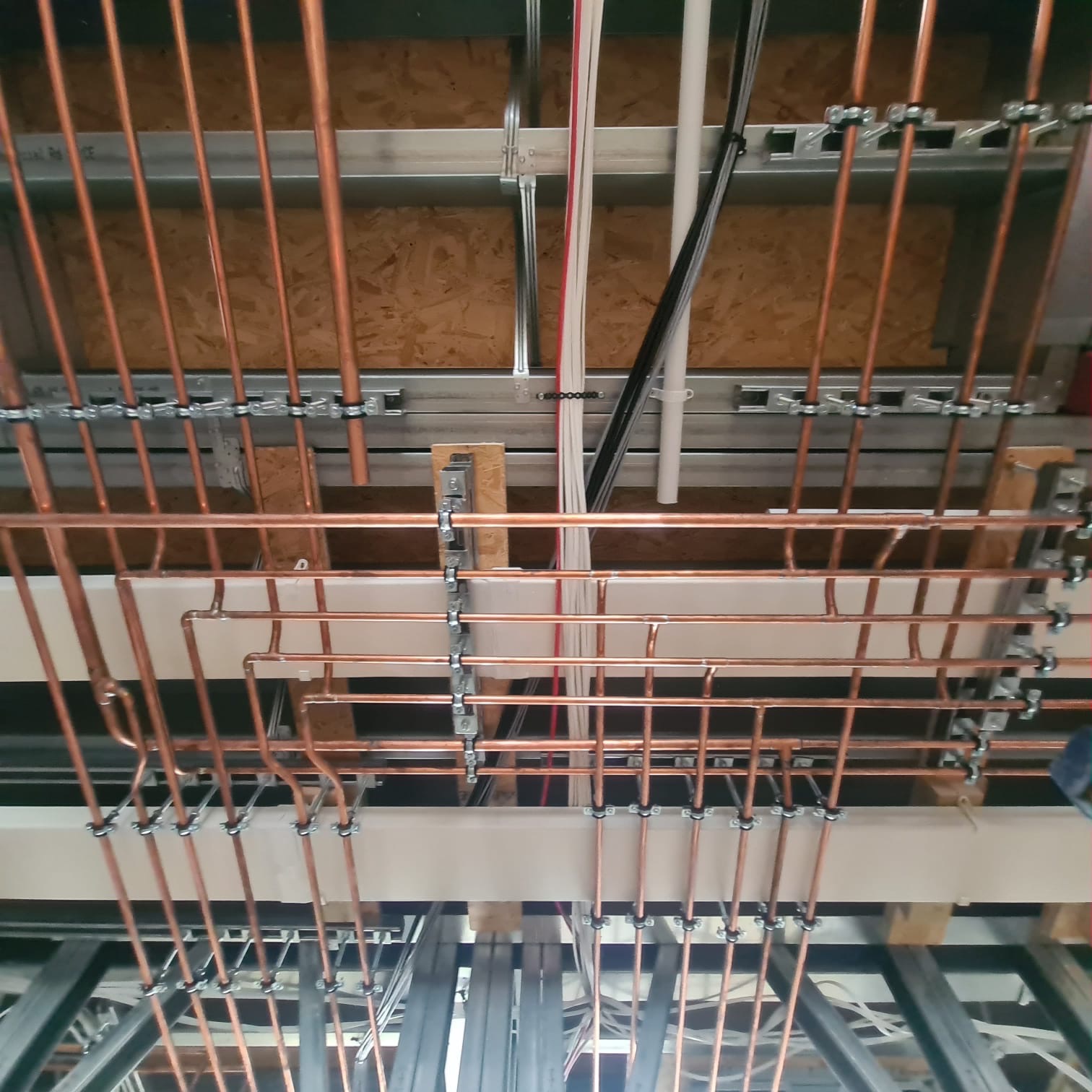 Miedziane rury instalacji hydraulicznej zamocowane pod sufitem, widoczne białe i czarne kable oraz metalowa konstrukcja wsporcza.