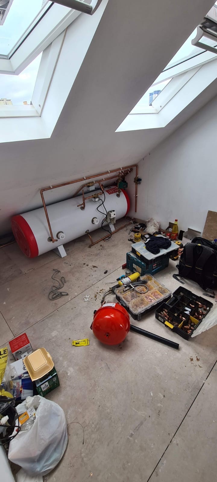 Montaż poziomego zbiornika ciepłej wody użytkowej w skośnym pomieszczeniu z oknami dachowymi, widoczne rury miedziane, narzędzia hydrauliczne i bałagan na podłodze.