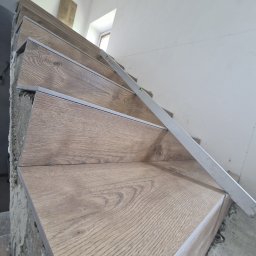 MAKS-FLIZ Uslugi Glazurnicze - Montaż drewnopodobnych płytek na schodach, poziomica w użyciu, surowy stan wykończenia ściany.