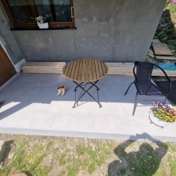 MAKS-FLIZ Uslugi Glazurnicze - Wyłożony jasnymi płytkami taras przed domem z drewnianym stołem, krzesłem z wikliny i doniczką z bratkami; w tle ułożone deski i kalosze.