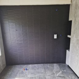 MAKS-FLIZ Uslugi Glazurnicze - Ściana łazienki wyłożona ciemnoszarą, heksagonalną mozaiką ceramiczną, z widoczną instalacją elektryczną i fragmentem półki wnękowej, oraz podłoga z szarych płytek imitujących marmur, z niebieskim...