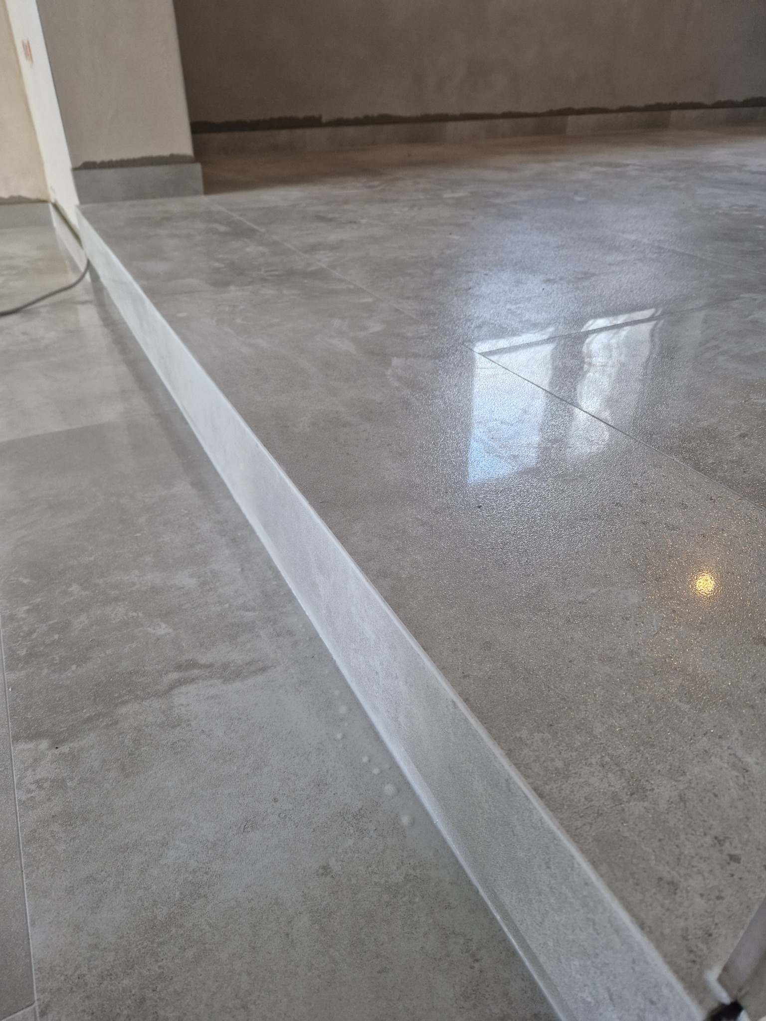 Ułożona, szara podłoga z płytek ceramicznych z widocznym progiem, w tle surowa ściana przygotowana do wykończenia.