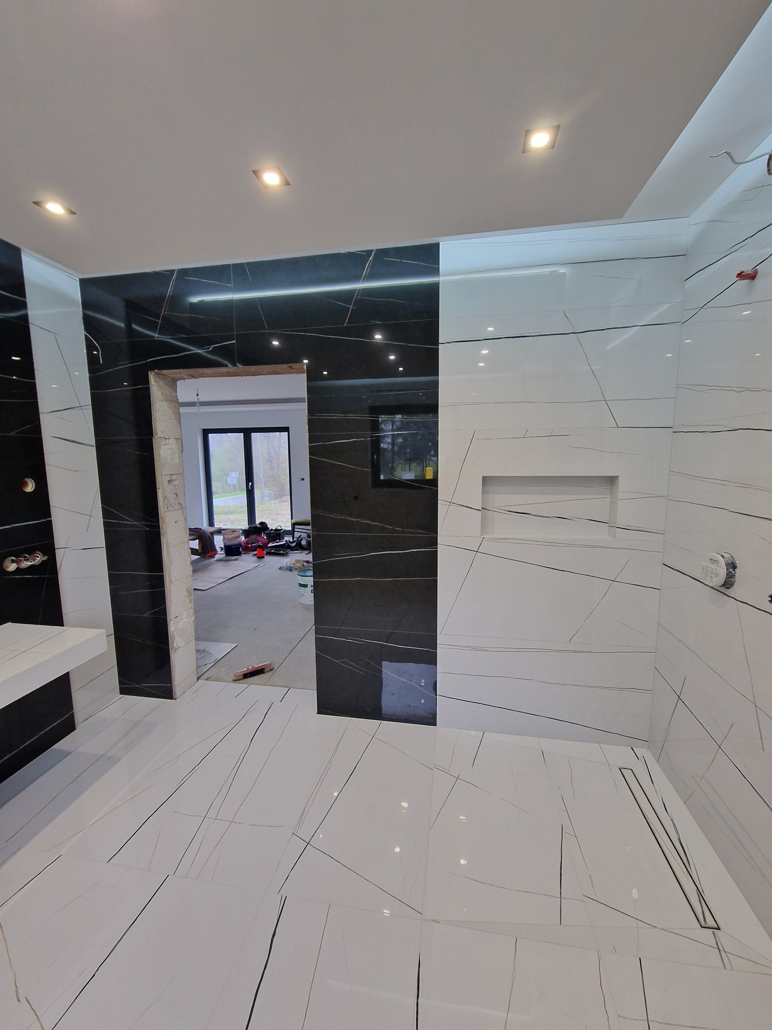 Nowoczesna łazienka w trakcie wykańczania: białe i czarne płytki imitujące marmur na ścianach i podłodze, widoczny odpływ liniowy w strefie prysznica, wnęka w ścianie, przez otwarty prześwit...