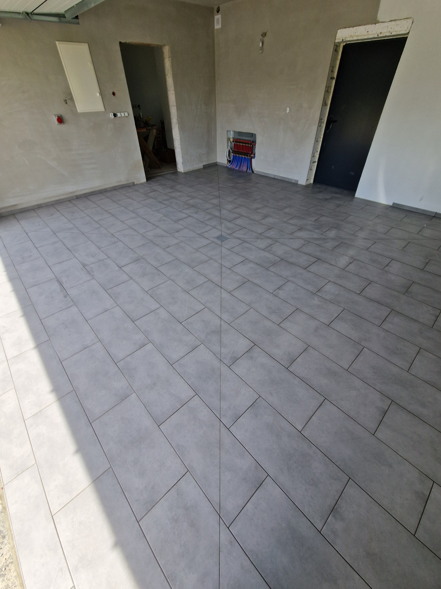 Ułożona, szara, prostokątna terakota w nowym pomieszczeniu. Widoczne drzwi i instalacja grzewcza w ścianie.