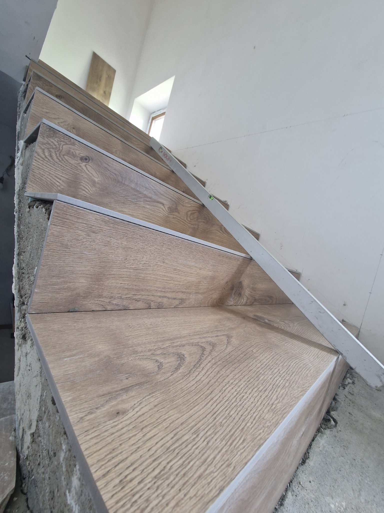 Montaż drewnopodobnych płytek na schodach, poziomica w użyciu, surowy stan wykończenia ściany.