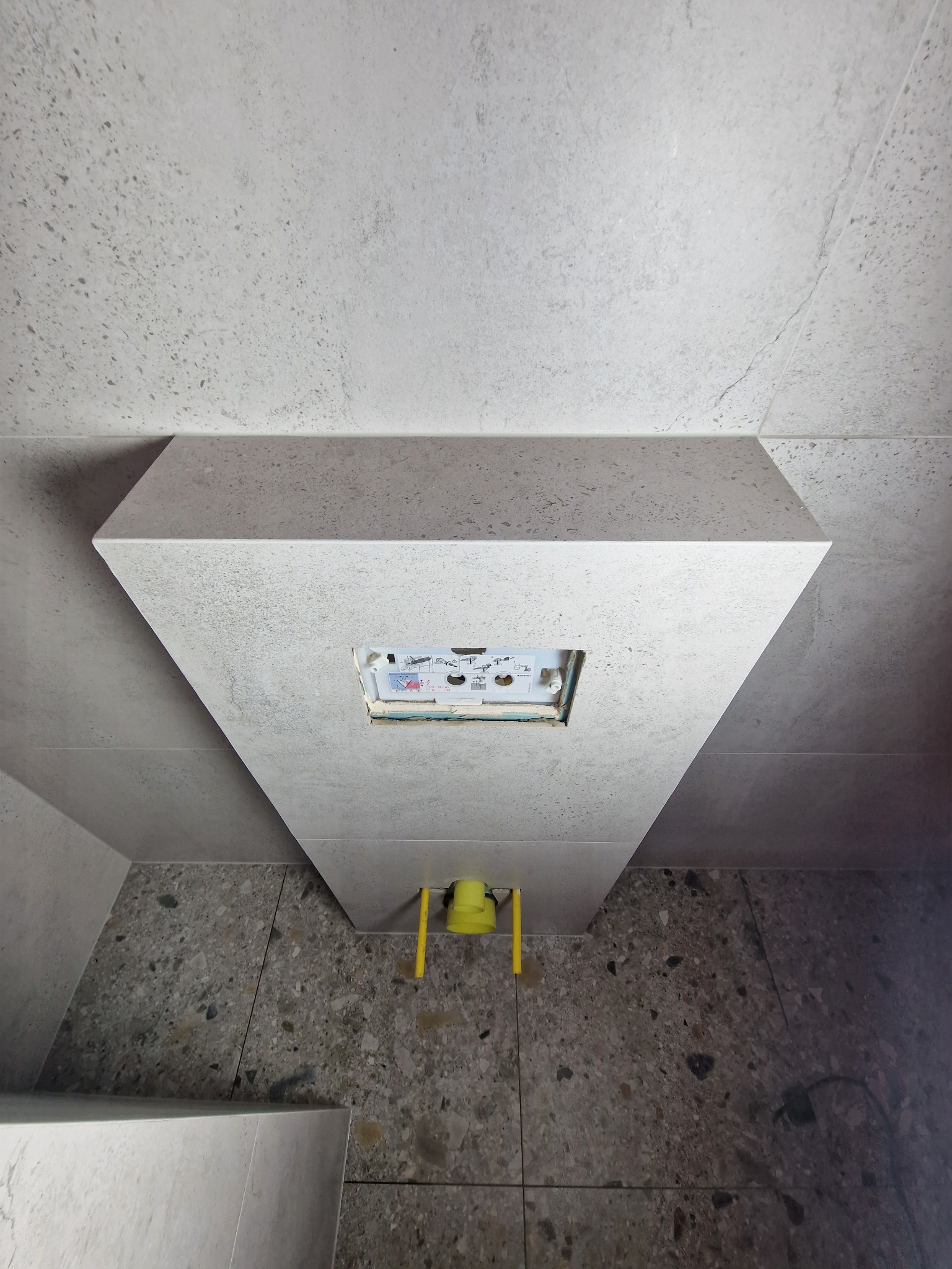 Obudowa stelaża podtynkowego WC wykończona szarymi płytkami imitującymi lastryko, widoczne żółte elementy instalacji hydraulicznej.