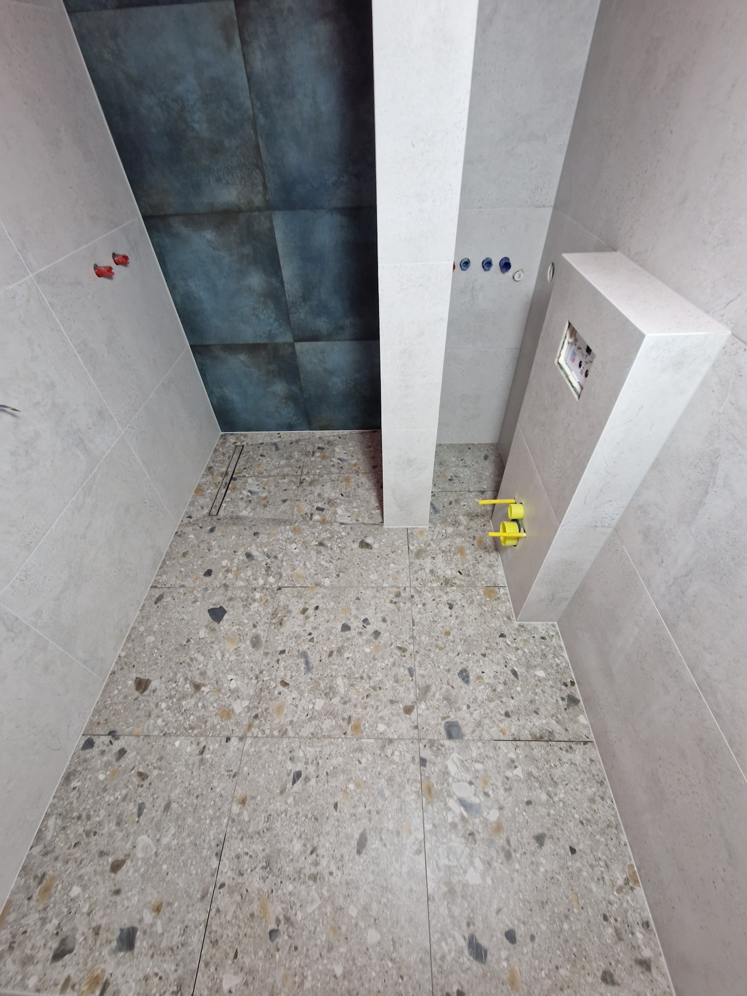 Łazienka w trakcie prac wykończeniowych: szare płytki podłogowe typu lastryko, częściowo ułożone szare płytki ścienne, widoczne elementy instalacji wodno-kanalizacyjnej, przymocowane plastikowe...