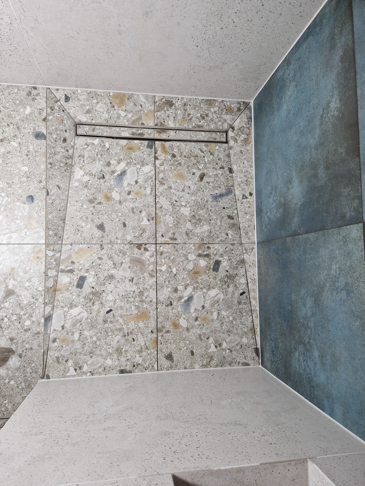 Nowoczesny prysznic z odpływem liniowym w posadzce, wykończony płytkami imitującymi lastryko i dużymi, błękitnymi płytami na ścianie. Widoczny fragment siedziska.