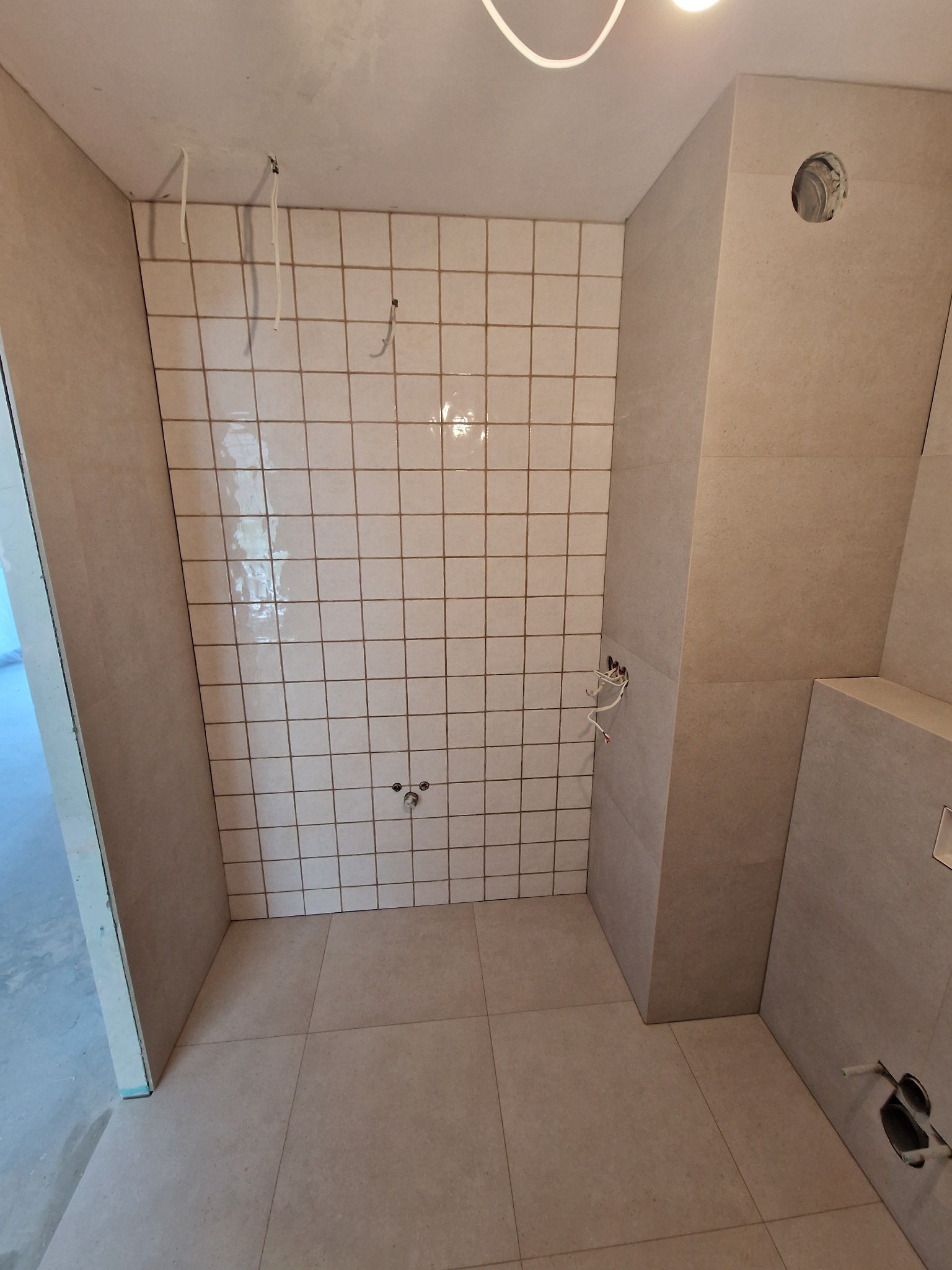 Łazienka w trakcie remontu: beżowe płytki ścienne i podłogowe ułożone w kwadraty, widoczne wystające przewody elektryczne i rury, brak armatury, surowy stan wykończenia.