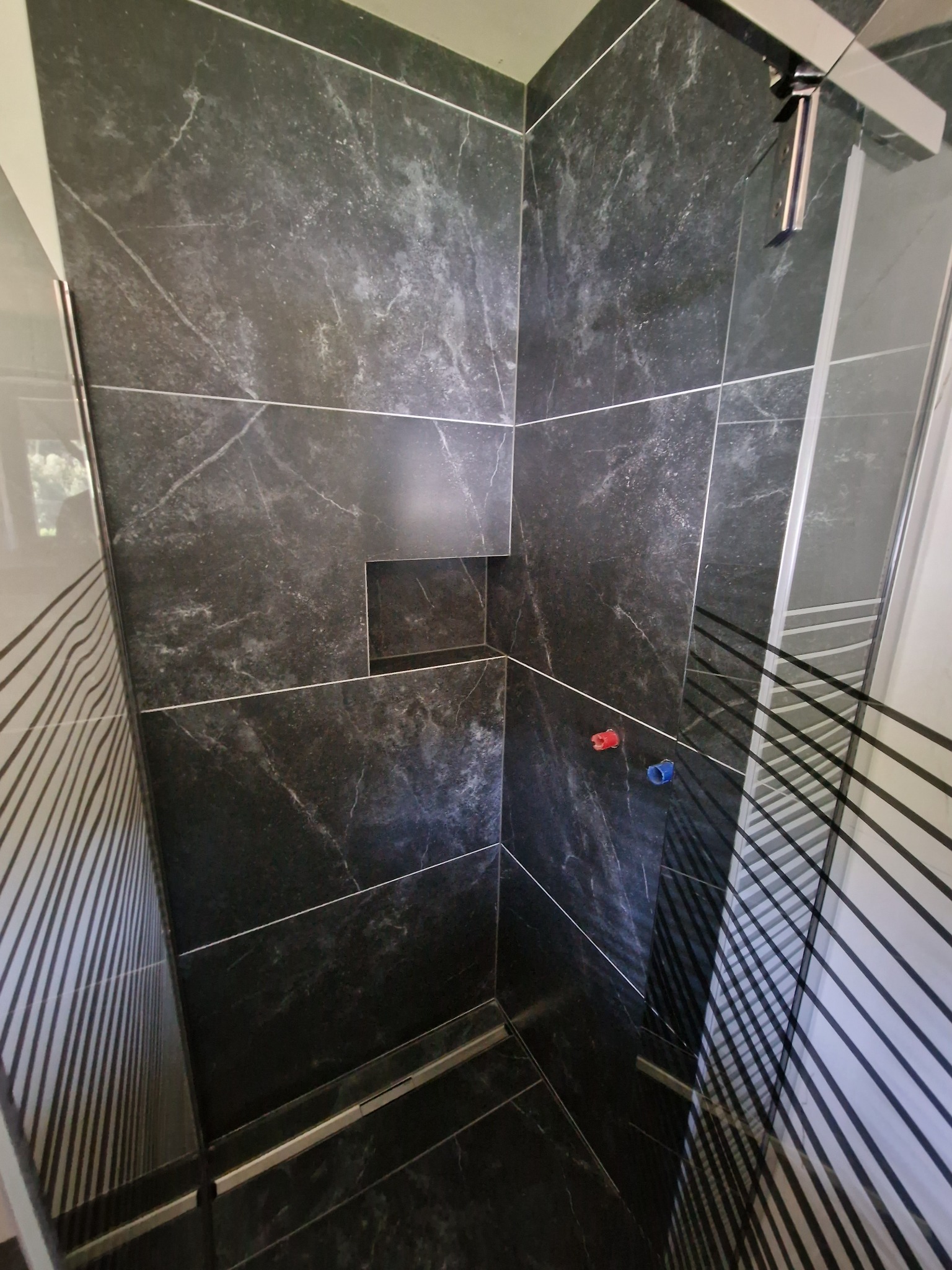 Kabina prysznicowa wyłożona czarnymi płytkami imitującymi marmur, z wnęką na kosmetyki i widocznymi przyłączami wody z oznaczeniami kolorystycznymi.