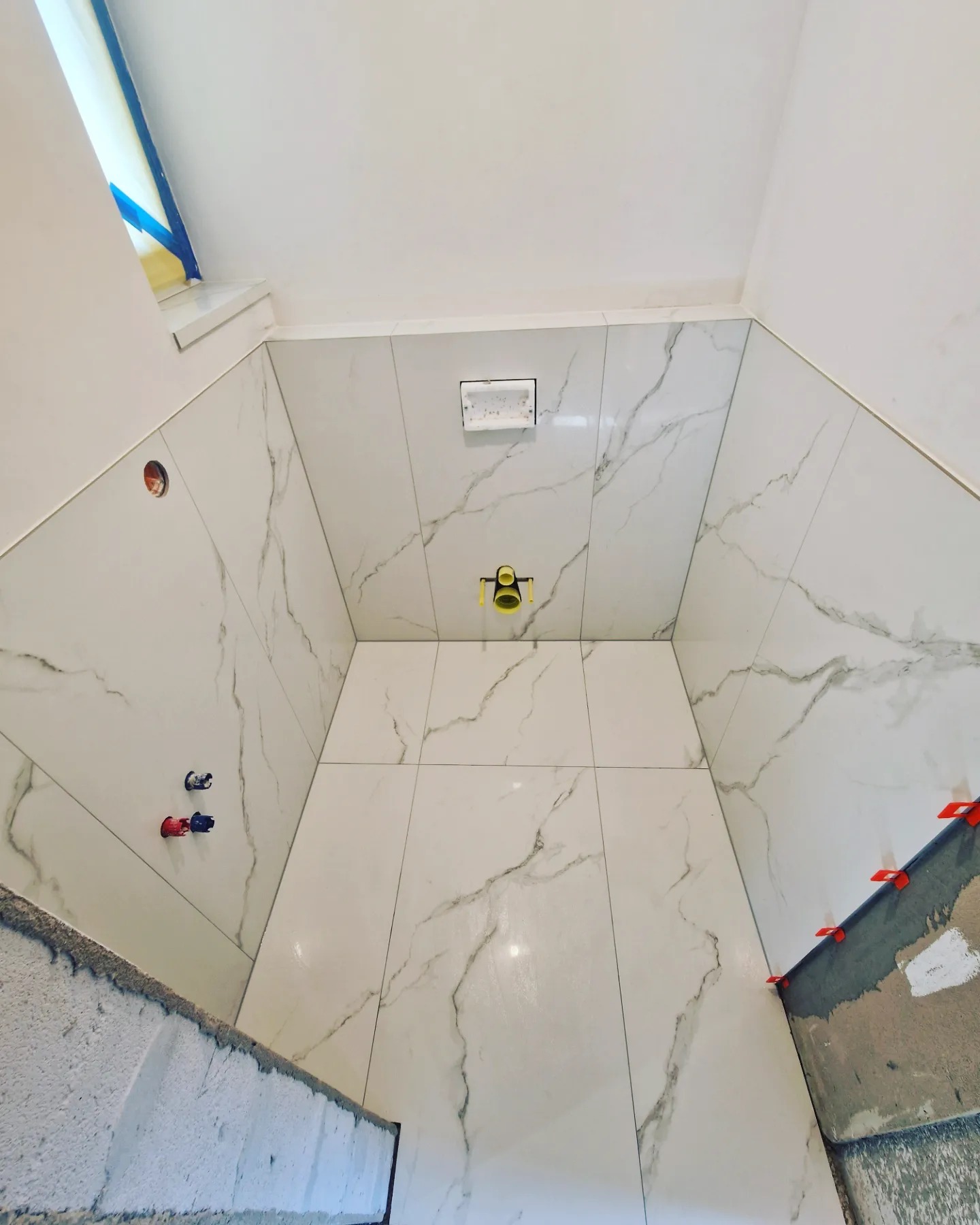 Montaż białych płytek imitujących marmur w narożnej kabinie prysznicowej, widoczne czerwone i niebieskie przyłącza wodne, żółty syfon podtynkowy i pomarańczowe krzyżyki dystansowe.
