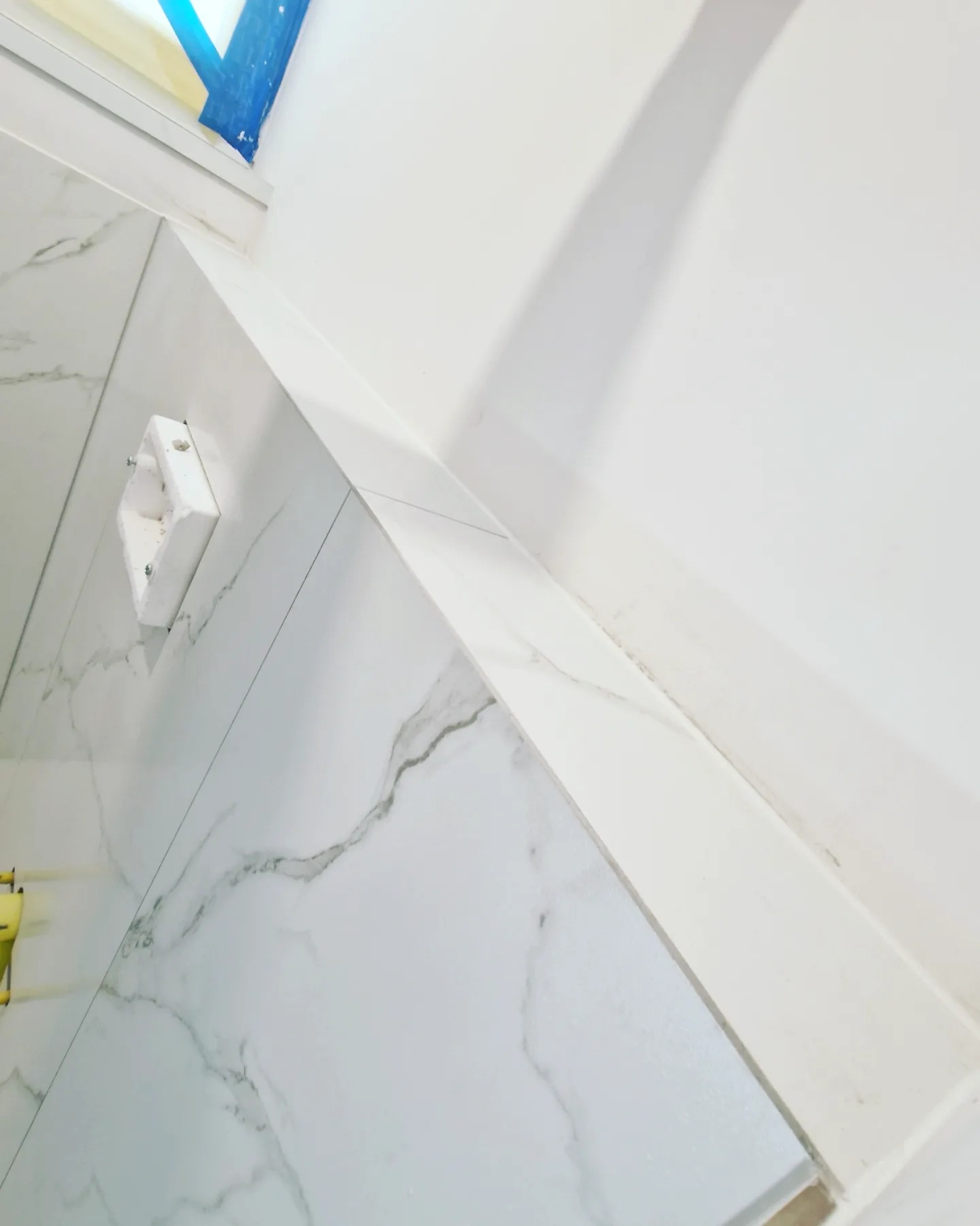 Ściana łazienki wyłożona białymi płytkami imitującymi marmur, z widoczną puszką elektryczną przed montażem osprzętu.