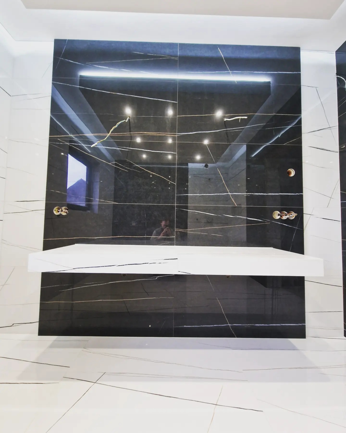 Wykończona ściana z czarnych płytek imitujących marmur z białymi żyłami i podświetleniem LED, uzupełniona białą półką w łazience w Rdzawce, widoczne przygotowane punkty instalacji wodnej.